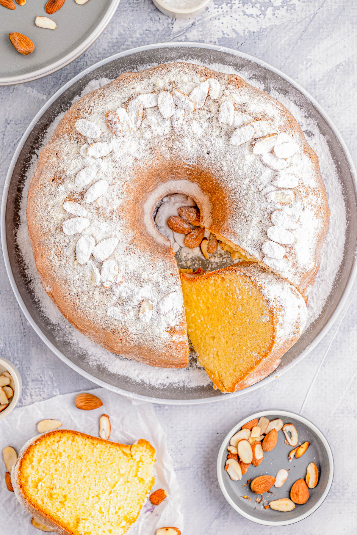 A bundt cake dusted with powdered sugar and topped with almonds. A slice is cut and placed on a plate. Additional almonds are scattered around.