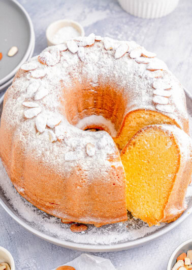 Bundt cake dusted with powdered sugar and topped with almond slices on a gray plate, featuring a single slice cut out; surrounded by scattered almonds and small dishes.