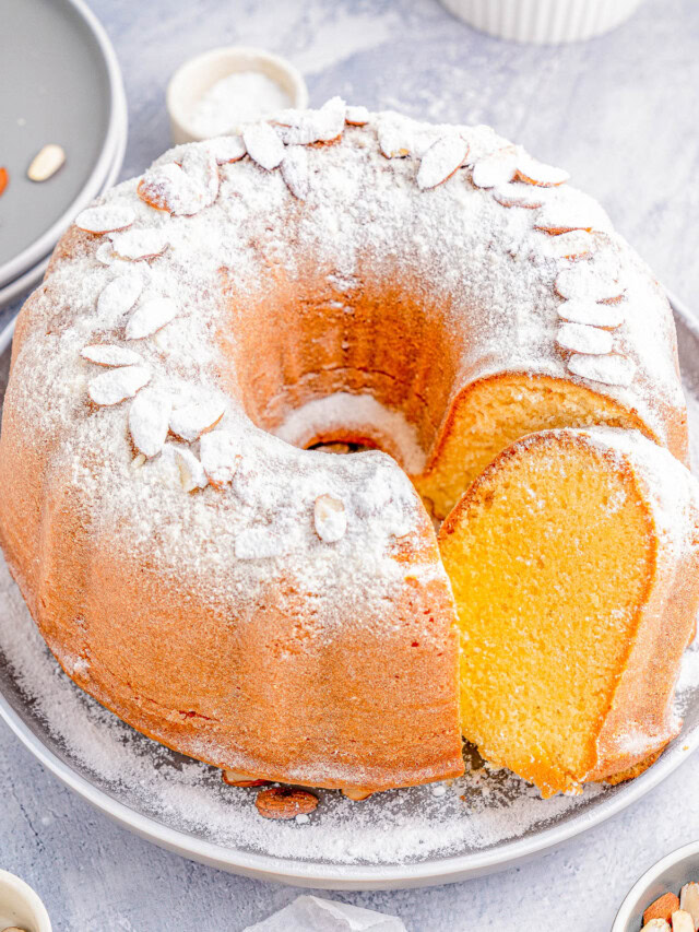 Bundt cake dusted with powdered sugar and topped with almond slices on a gray plate, featuring a single slice cut out; surrounded by scattered almonds and small dishes.