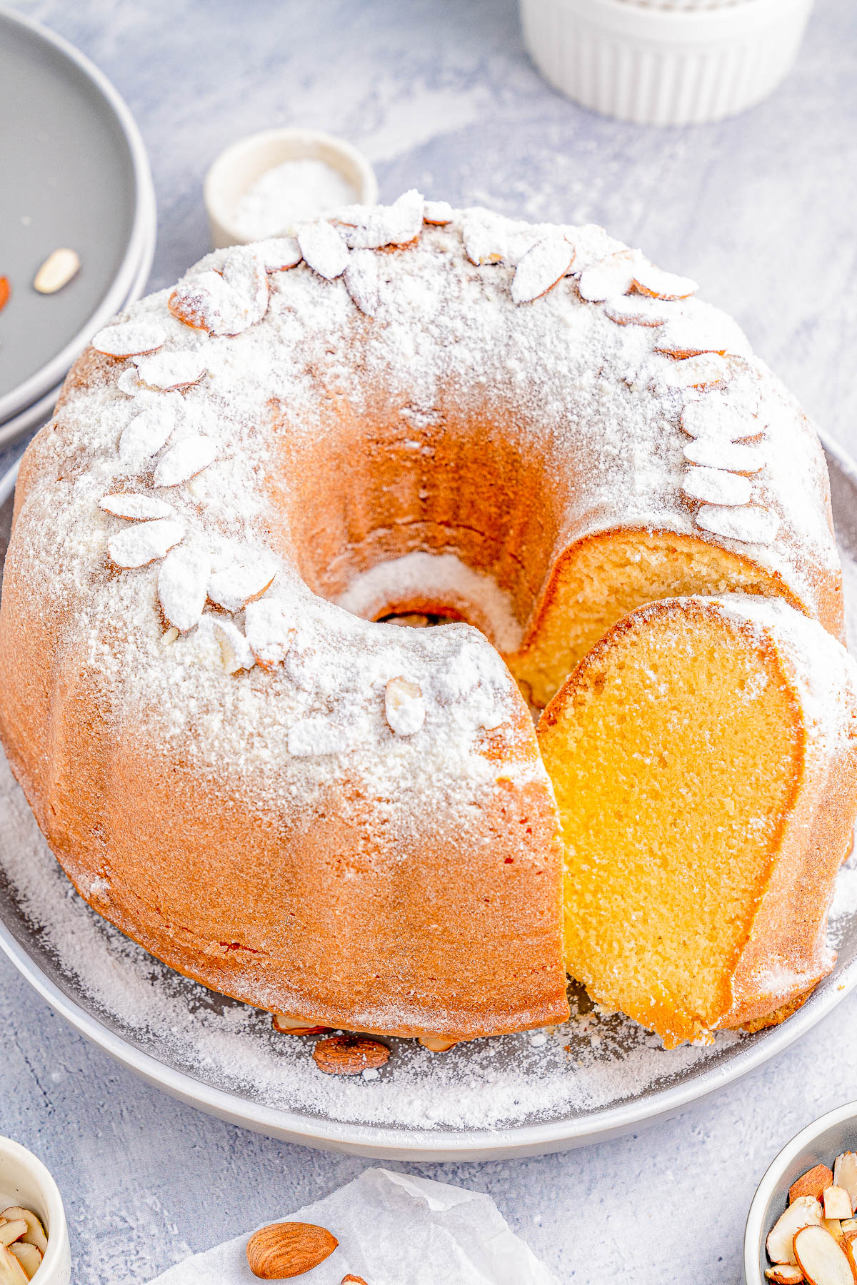 Bundt cake dusted with powdered sugar and topped with almond slices on a gray plate, featuring a single slice cut out; surrounded by scattered almonds and small dishes.