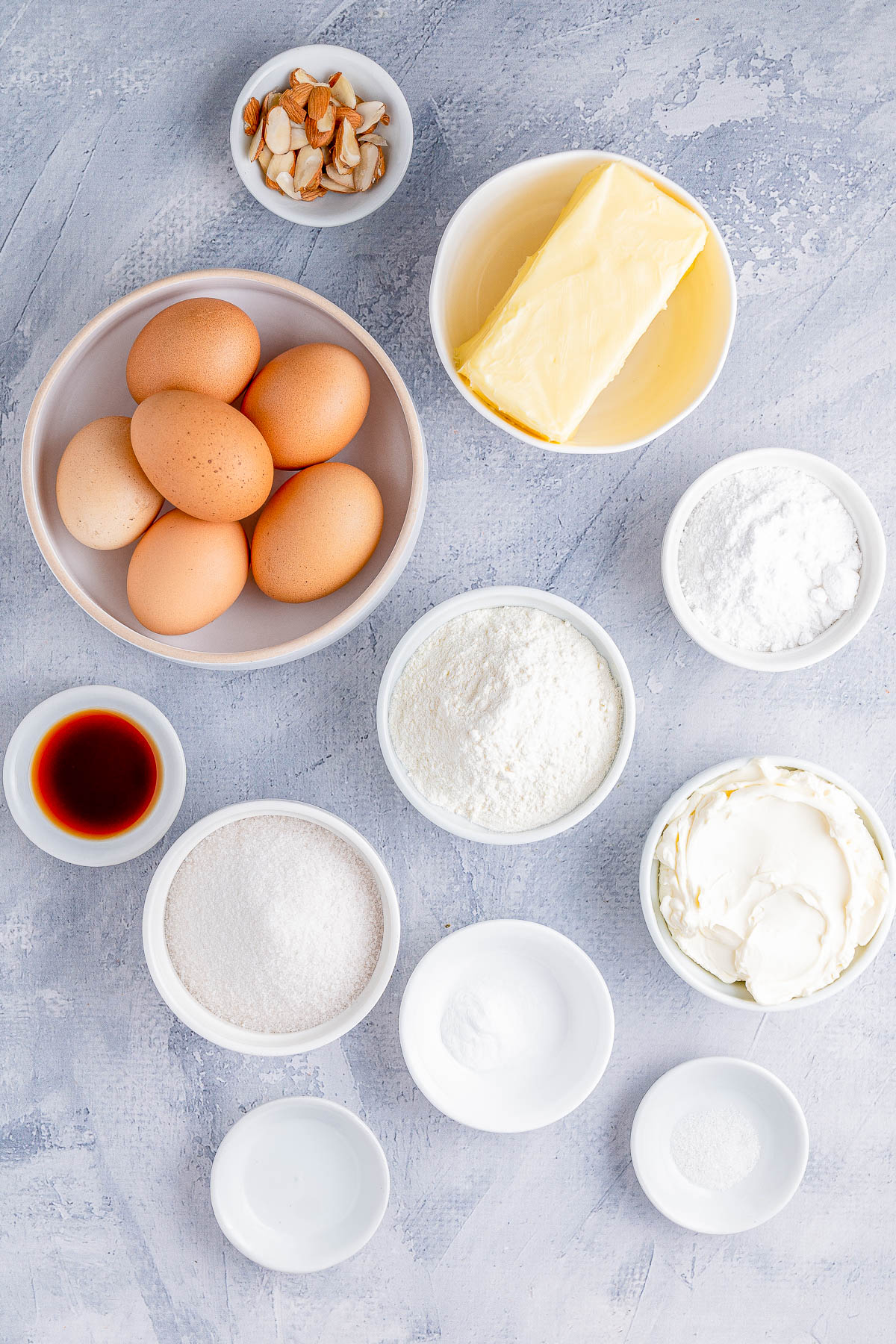 Ingredients for baking including eggs, butter, flour, sugar, vanilla extract, baking powder, baking soda, sour cream, and sliced almonds arranged on a textured surface.