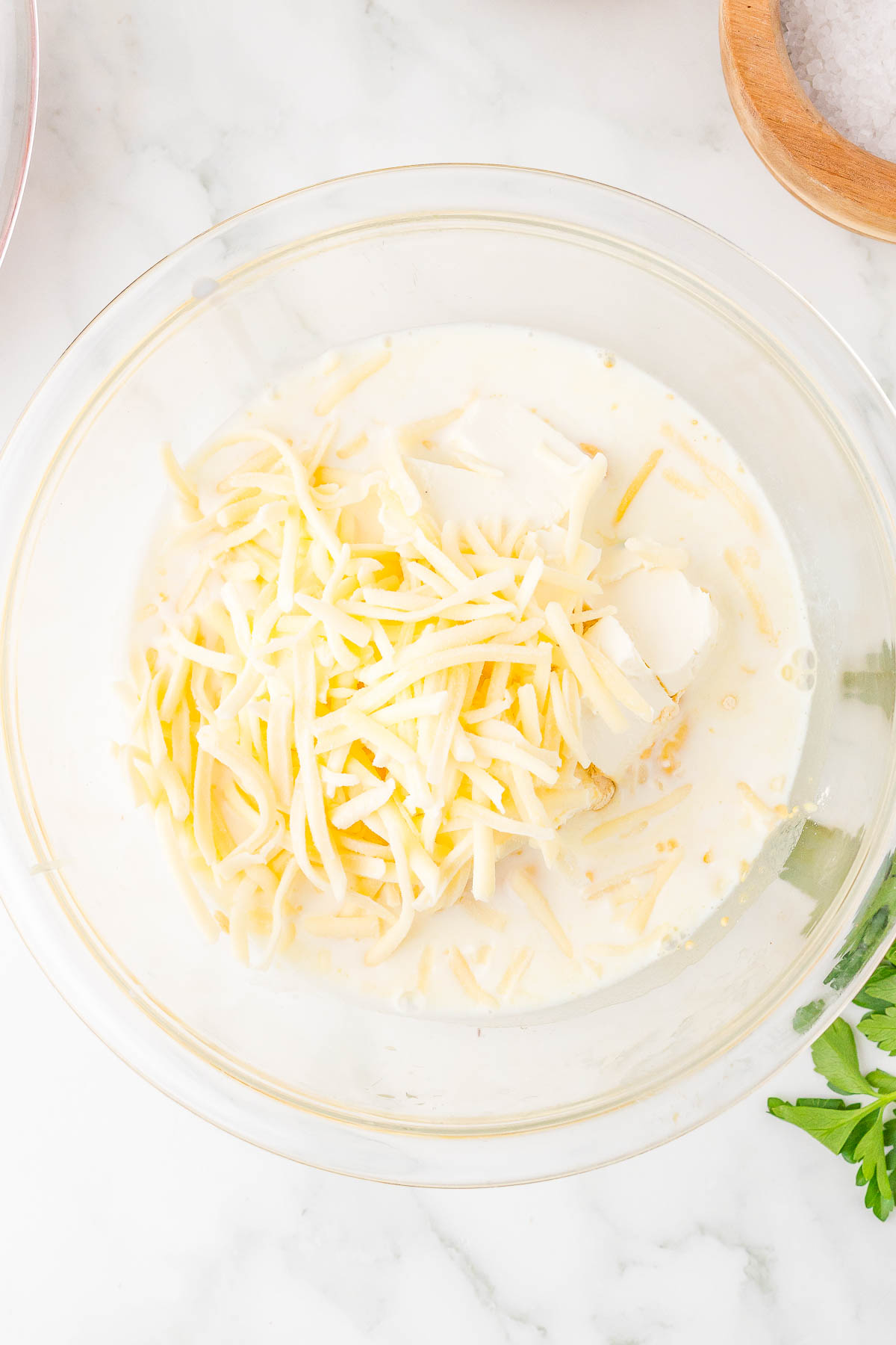 A glass bowl with shredded cheese and creamy white liquid on a marble countertop.