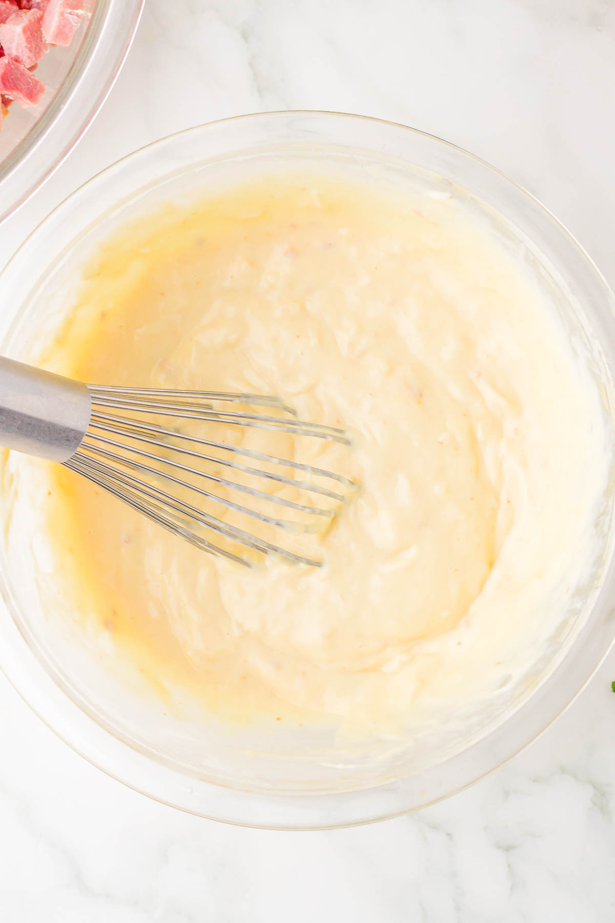 A bowl of thick, creamy batter is being mixed with a metal whisk on a marble countertop.