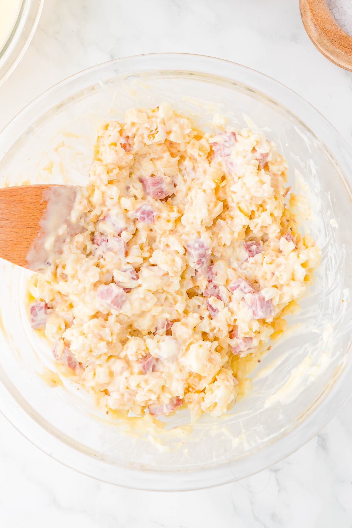 A glass bowl contains a mixture of chopped ham, shredded cheese, and mayonnaise, stirred with a wooden spatula.