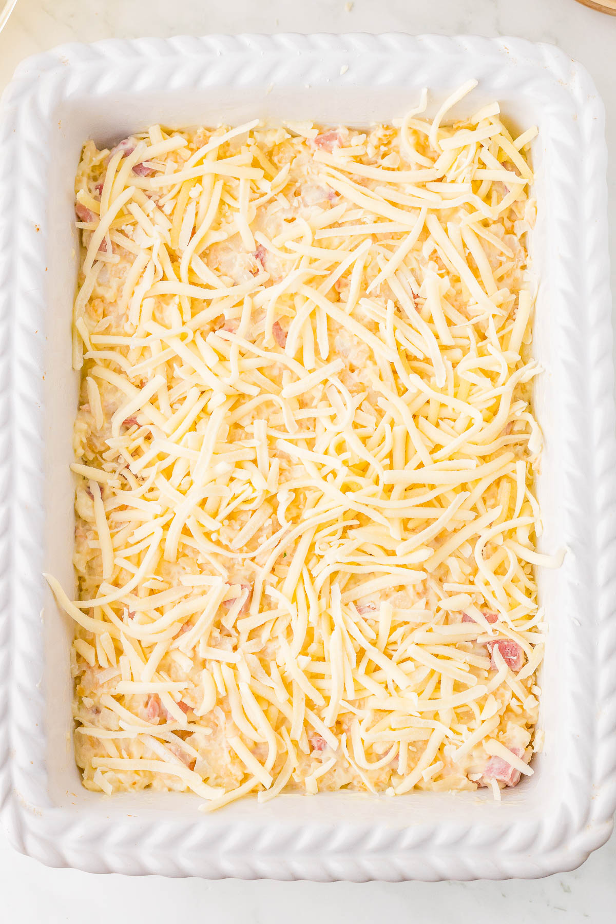 A rectangular baking dish filled with a cheese-topped mixture containing chopped ingredients, ready to be baked.