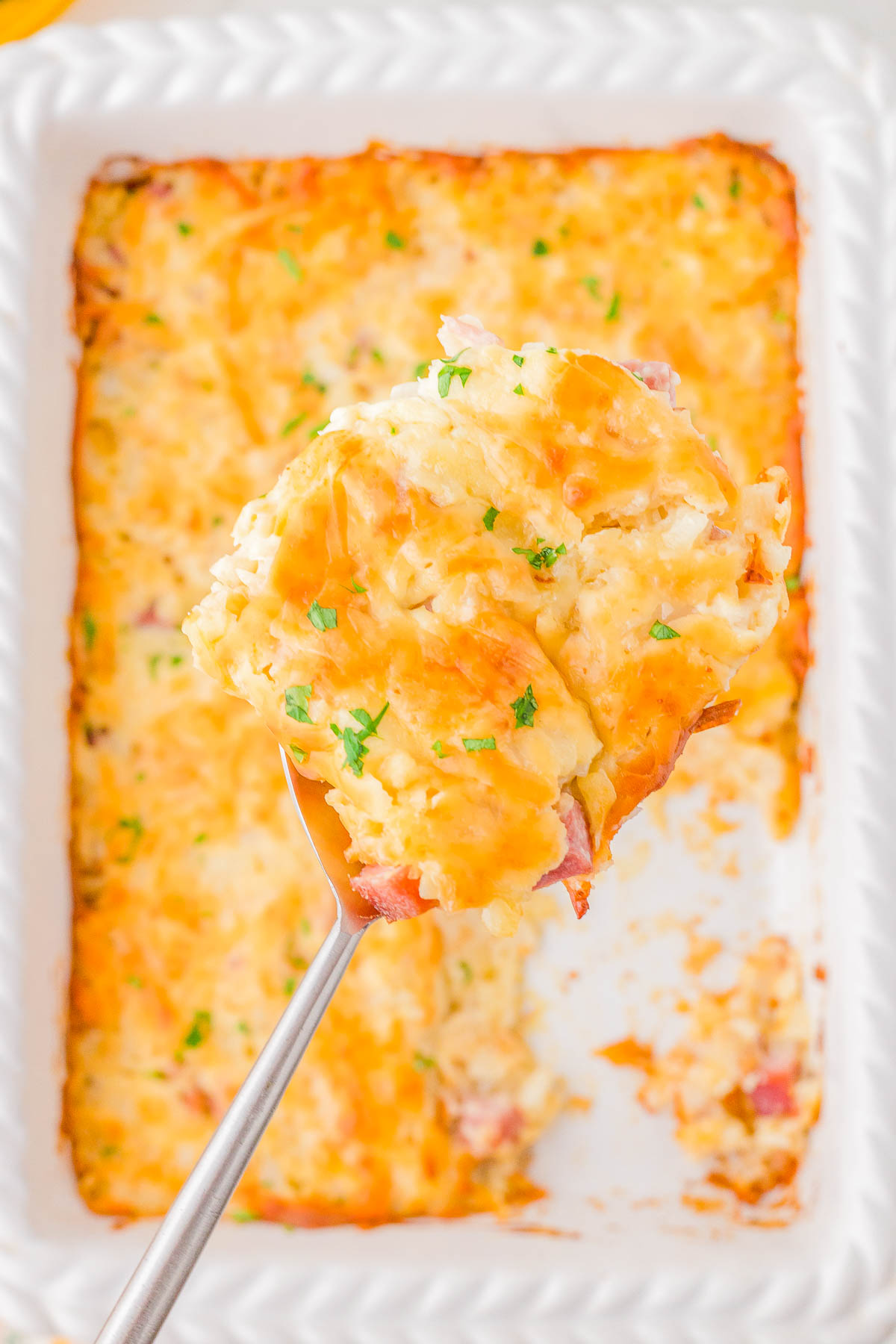 A close-up of a serving spoon lifting a portion of cheesy casserole with a golden-brown top, garnished with herbs, from a rectangular white baking dish.