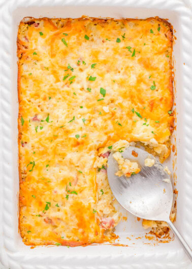 A baked casserole in a white dish, topped with melted cheese and garnished with herbs. A large serving spoon rests in an empty section.