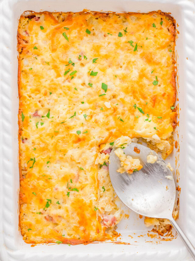 A baked casserole in a white dish, topped with melted cheese and garnished with herbs. A large serving spoon rests in an empty section.