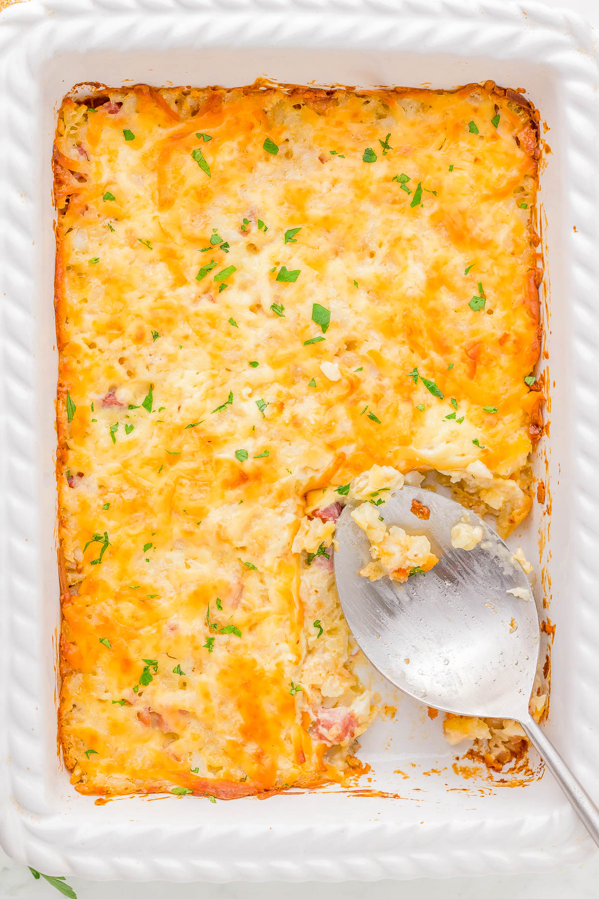 A baked casserole in a white dish, topped with melted cheese and garnished with herbs. A large serving spoon rests in an empty section.