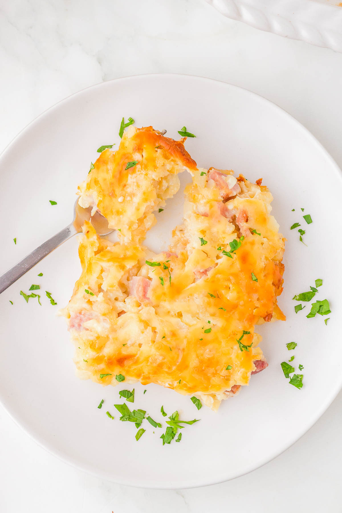 A serving of cheesy baked casserole with diced ham and garnished with chopped parsley on a white plate. A fork rests nearby.