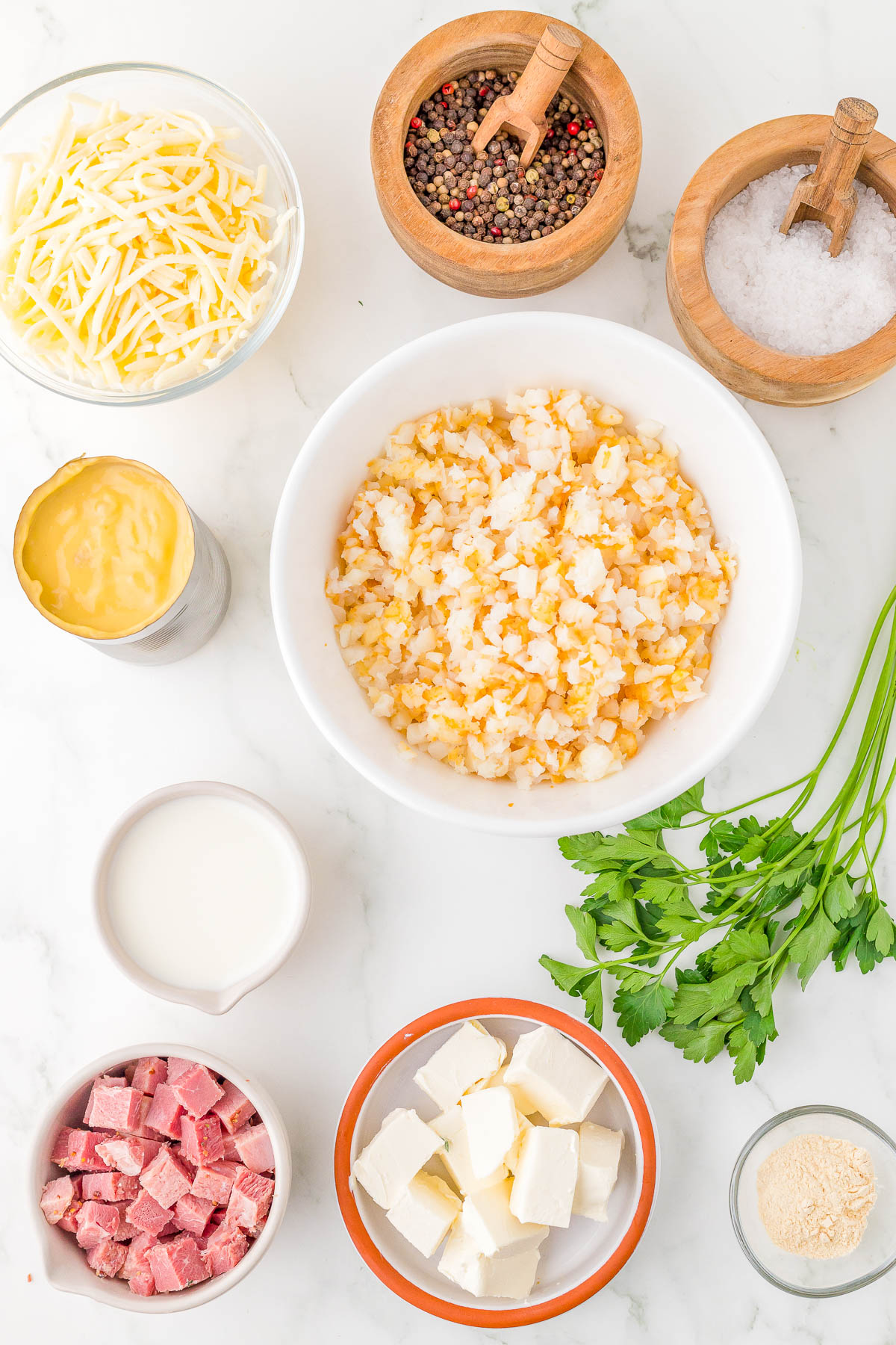 Ingredients arranged on a countertop include shredded cheese, pepper, salt, creamed corn, milk, diced ham, cream cheese cubes, parsley, hash browns, and garlic powder.