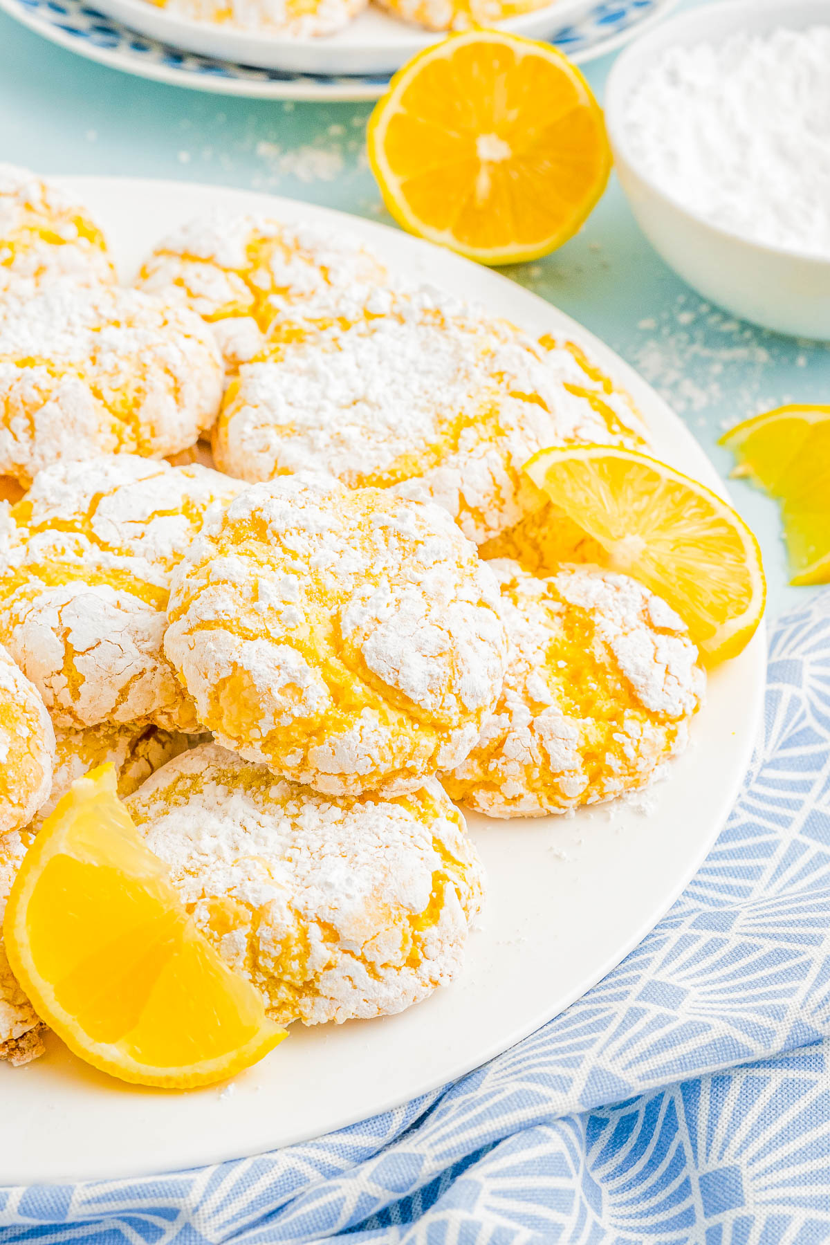 Plate of powdered sugar-coated lemon cookies with lemon slices on a blue patterned cloth.