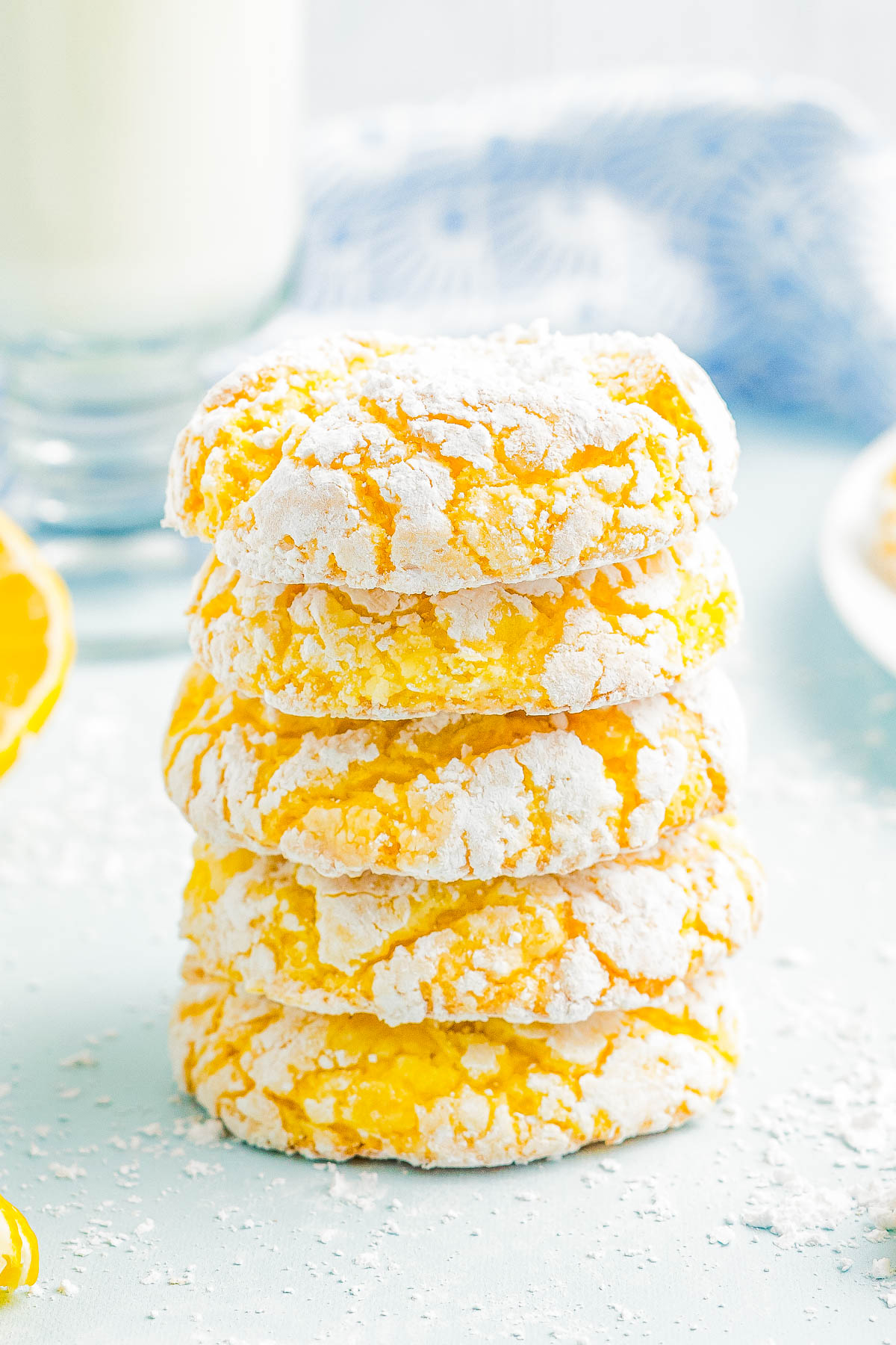 A stack of five lemon crinkle cookies dusted with powdered sugar is placed on a light blue surface. A glass of milk and a partially visible lemon are in the background.