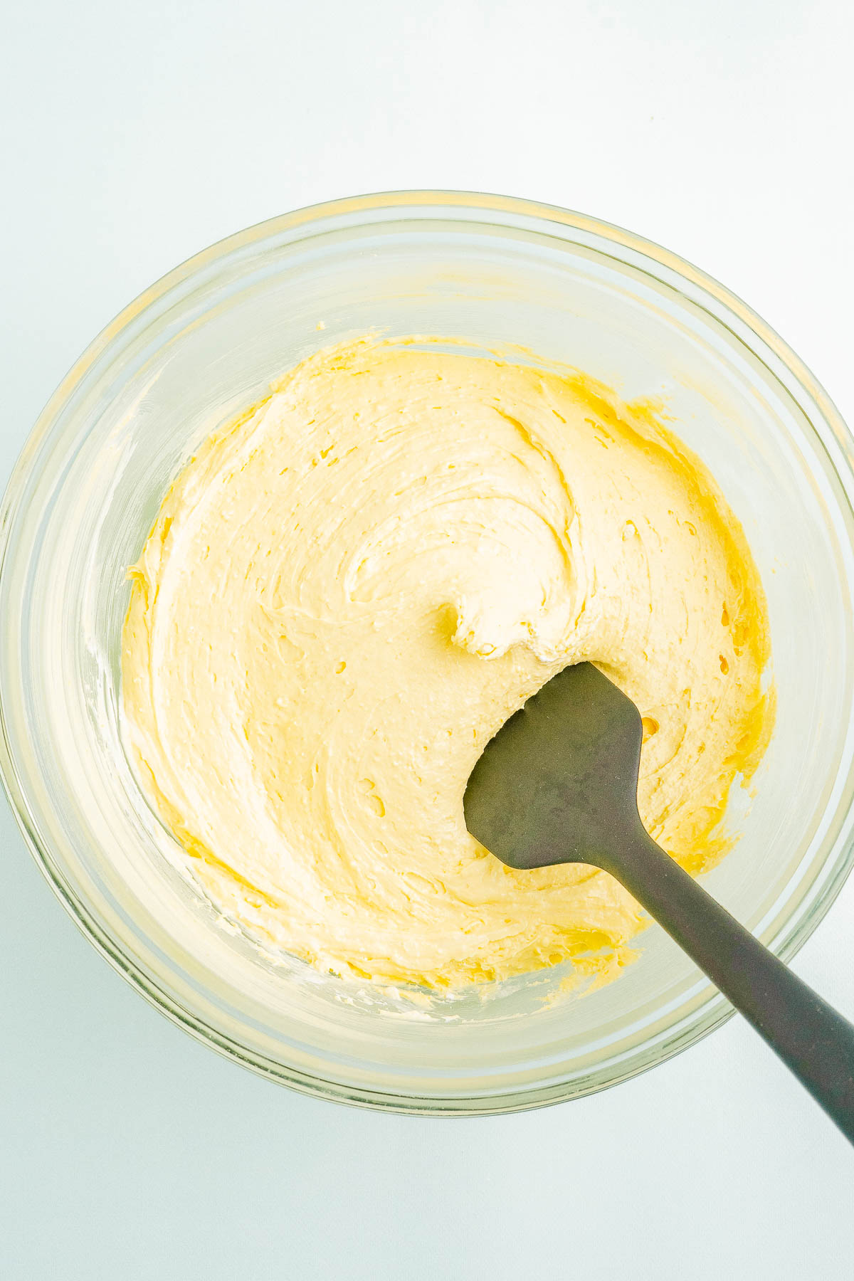 A glass bowl with creamy yellow cake batter being mixed by a black spatula.