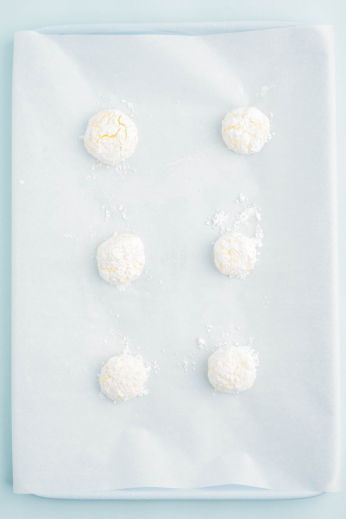 Six white powdered dough balls are spaced evenly on a sheet of parchment paper.