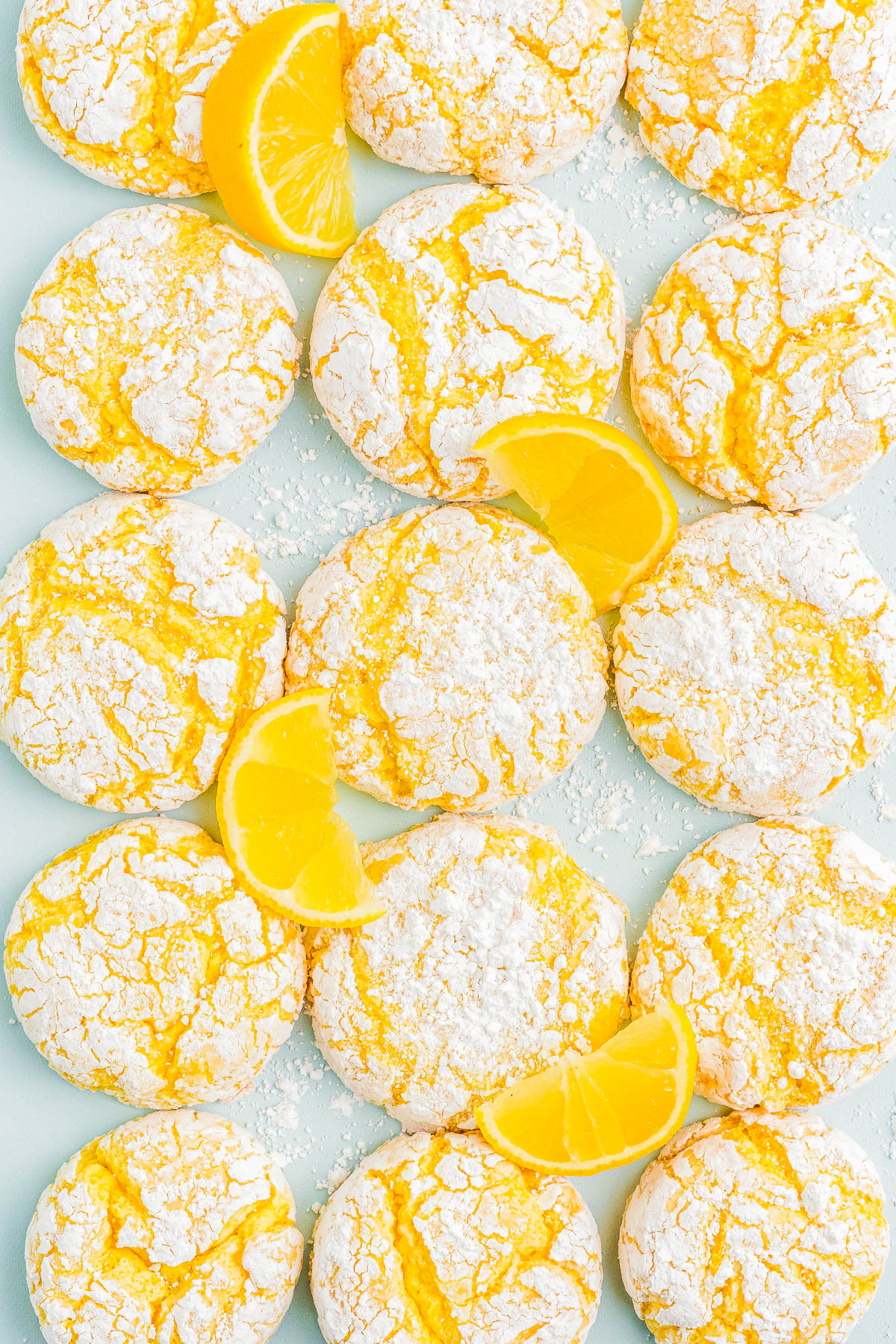 Yellow crinkle cookies dusted with powdered sugar, accompanied by lemon slices on a light blue background.