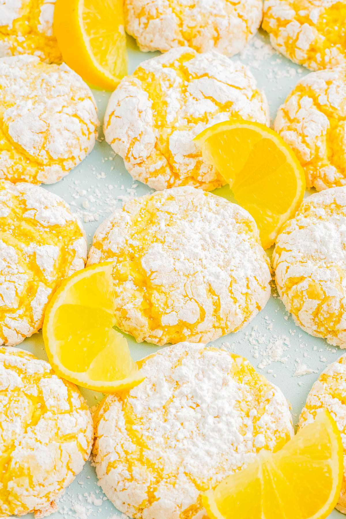 Yellow crinkle cookies dusted with powdered sugar, surrounded by fresh lemon wedges on a light background.