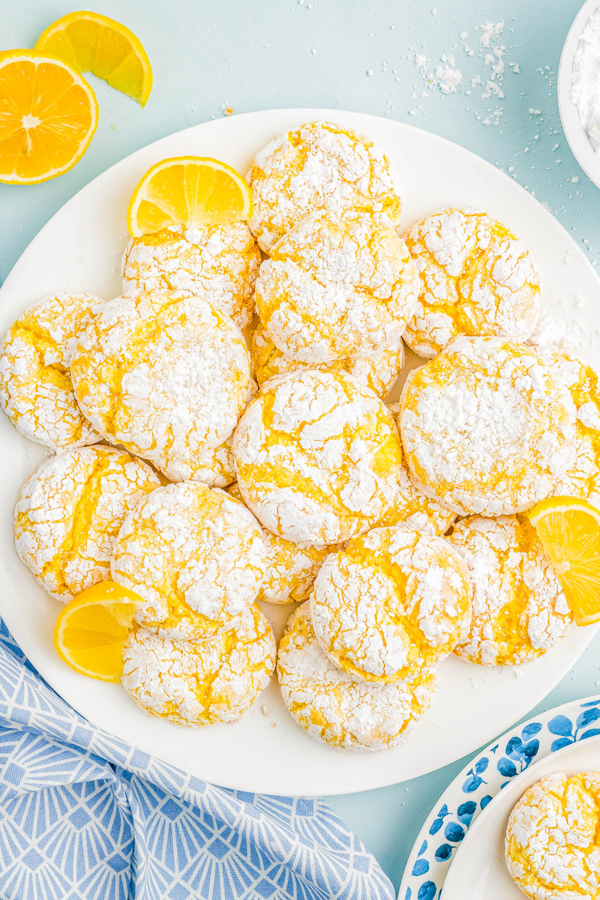 A plate of lemon crinkle cookies dusted with powdered sugar, garnished with lemon slices, placed on a light blue surface near a patterned cloth.