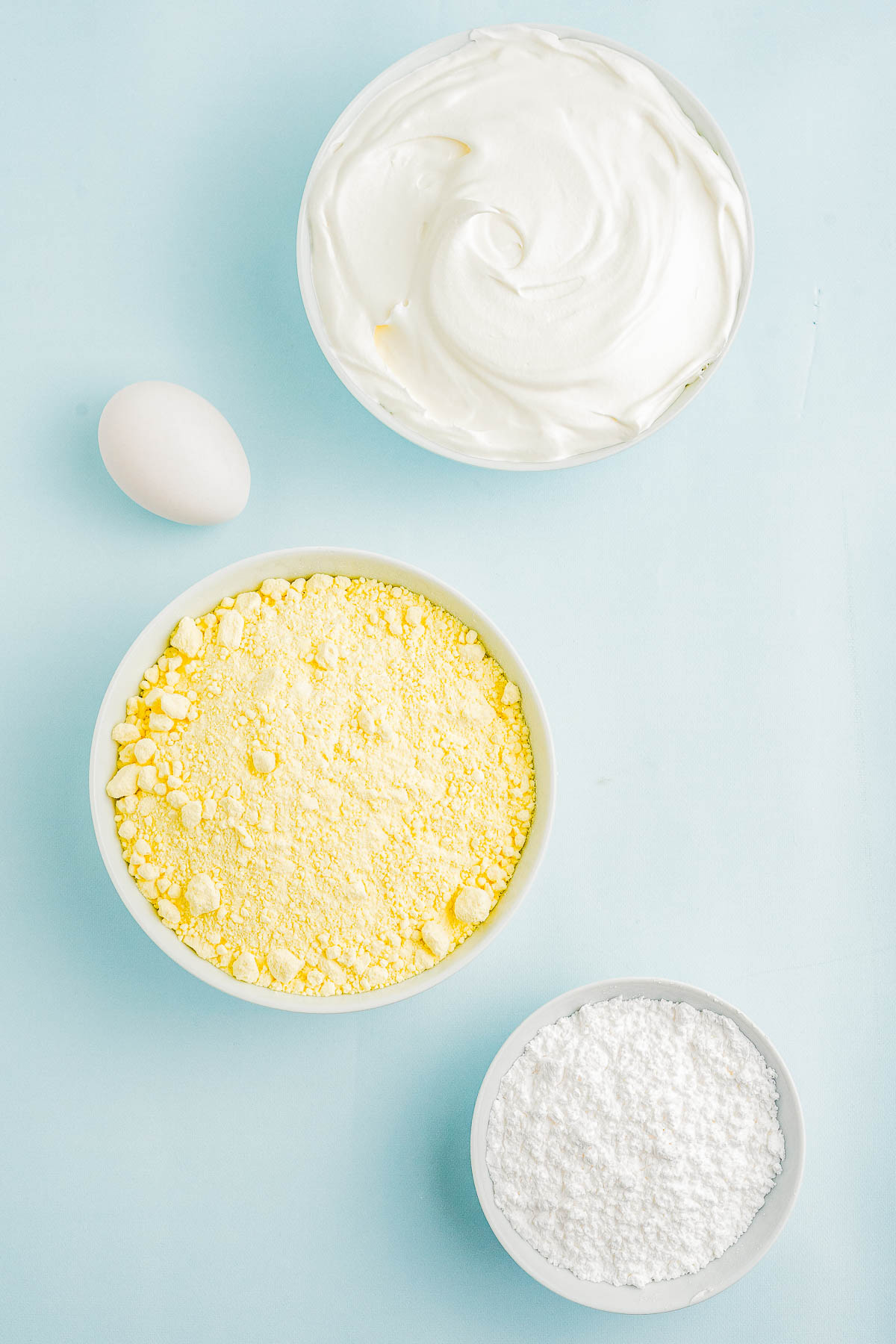 Bowls of whipped cream, yellow powder, and white powder are arranged on a light blue surface with a whole egg.