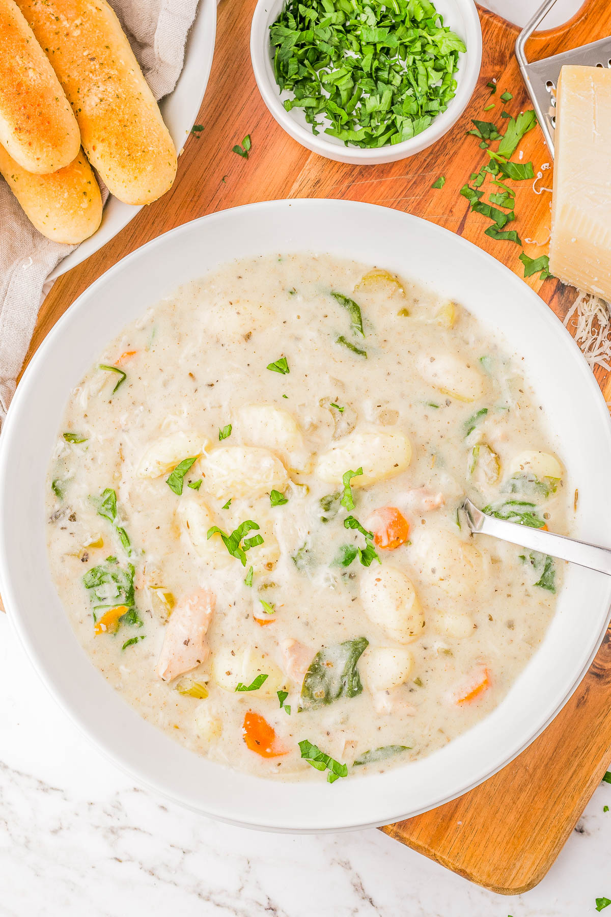 A bowl of creamy soup with gnocchi, spinach, and carrots, topped with chopped herbs. A spoon rests in the soup. Breadsticks and a dish of chopped herbs are on the side.