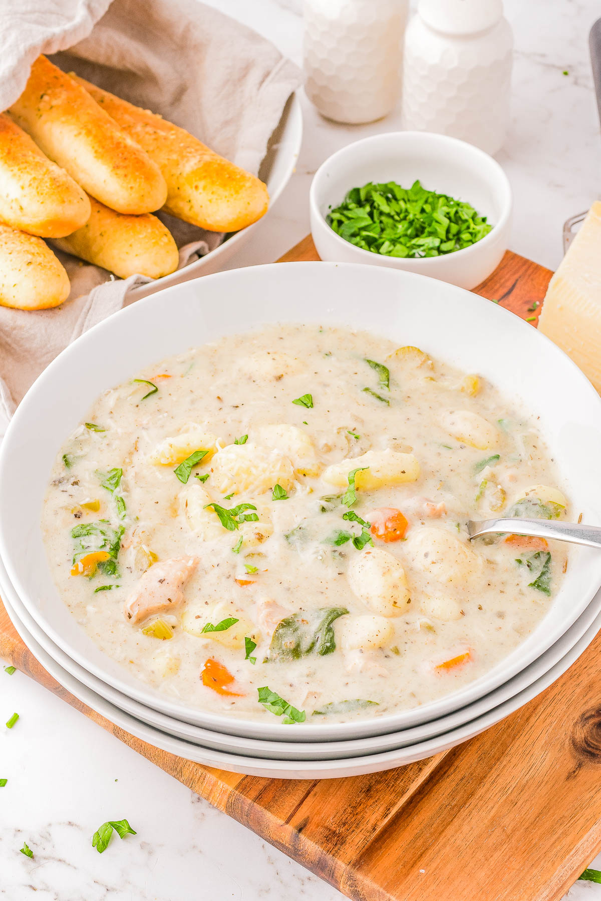 A bowl of creamy soup with vegetables, herbs, and gnocchi, garnished with chopped parsley. Breadsticks are in the background on a linen napkin.