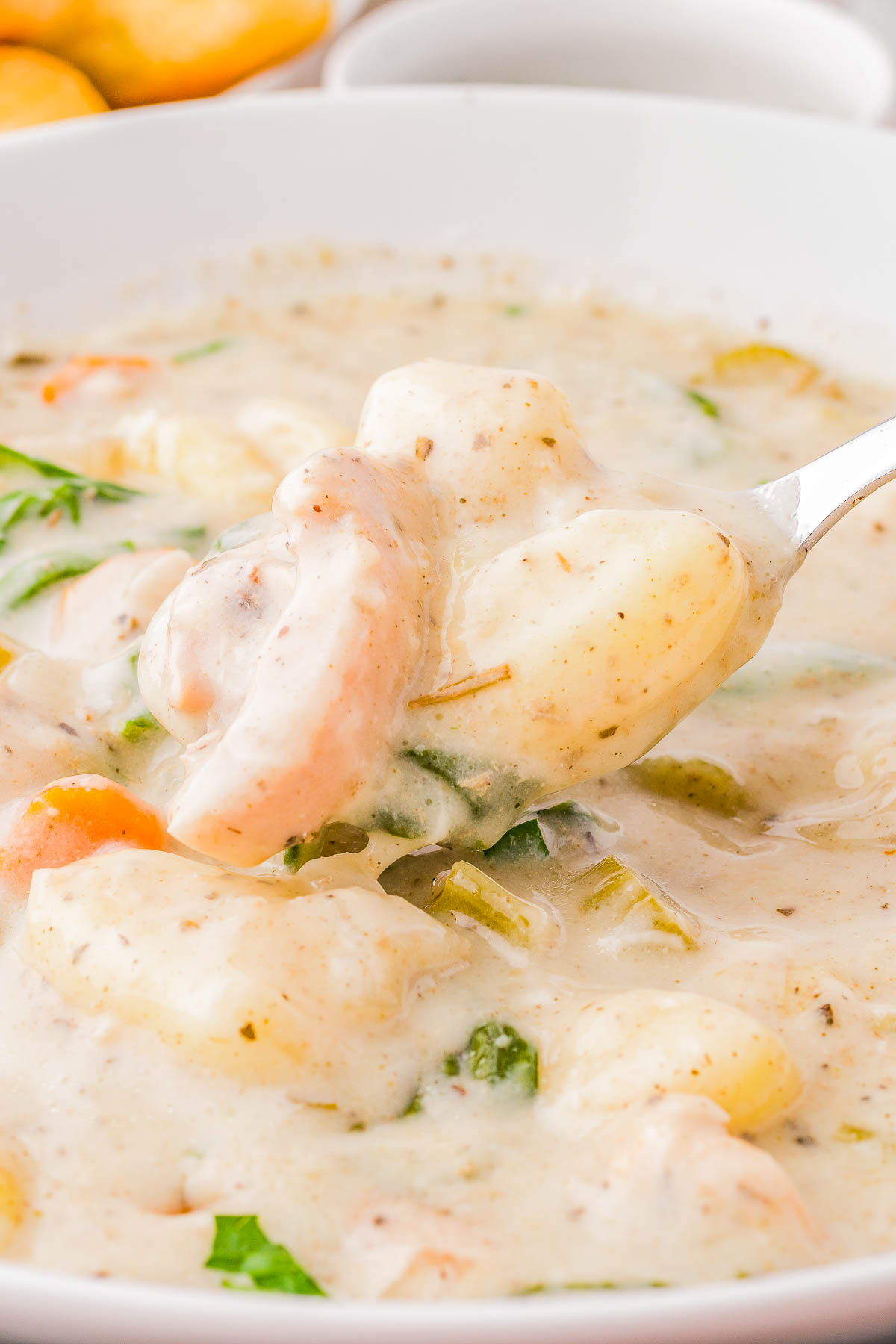 A bowl of creamy seafood chowder with shrimp, potatoes, carrots, and green herbs. A spoon is lifting a portion.