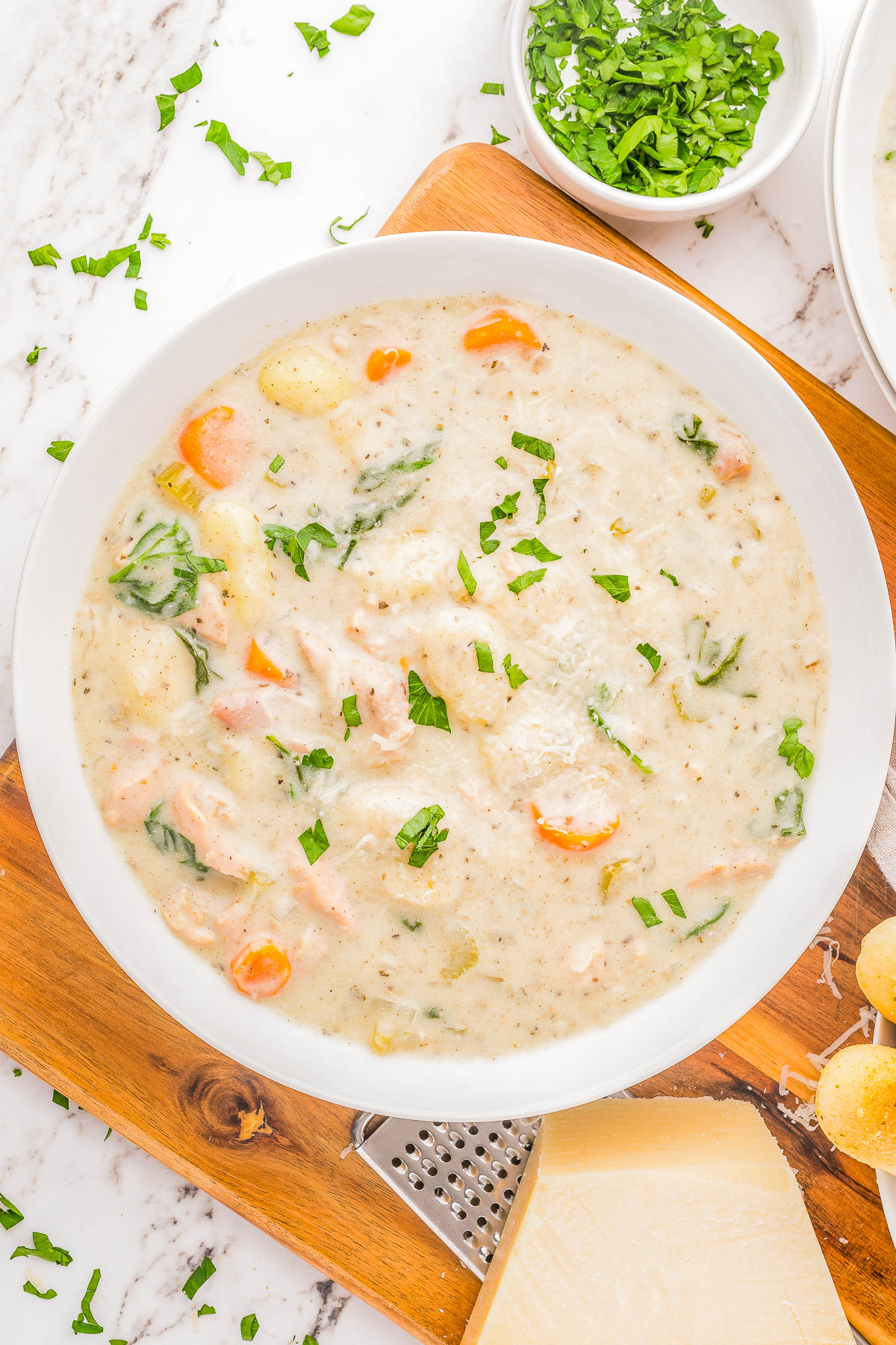 Bowl of creamy chicken gnocchi soup with carrots and spinach, garnished with parsley, on a wooden board next to grated cheese and a bowl of chopped herbs.