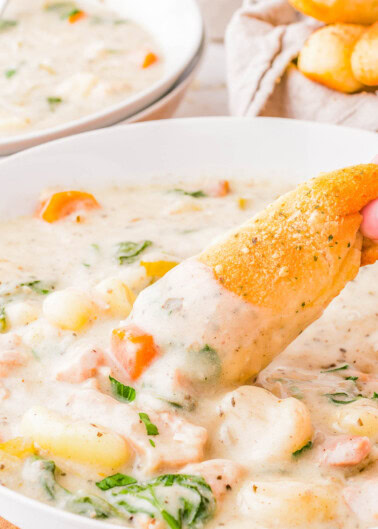 A bowl of creamy chicken gnocchi soup with visible chunks of chicken, carrots, and spinach. A hand dips a piece of bread into the soup. Another bowl and bread are blurred in the background.