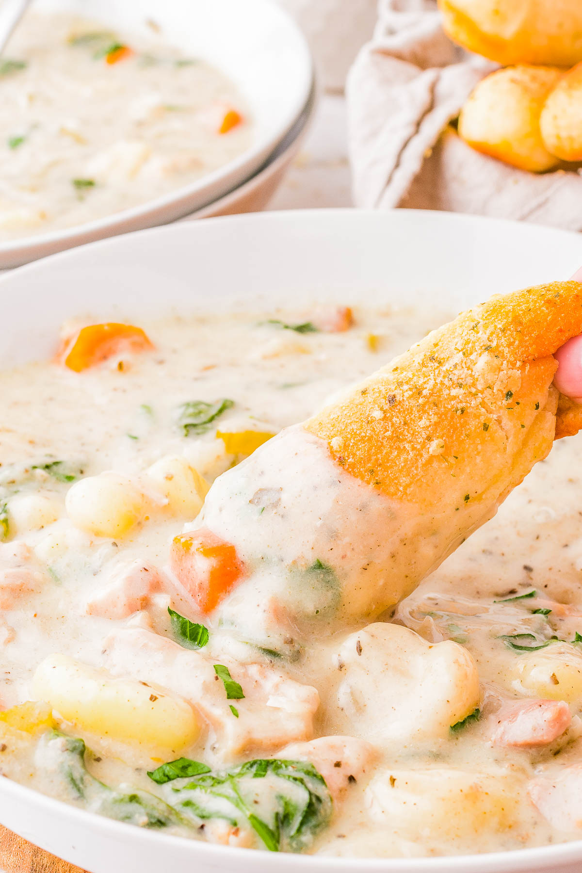 A bowl of creamy chicken gnocchi soup with visible chunks of chicken, carrots, and spinach. A hand dips a piece of bread into the soup. Another bowl and bread are blurred in the background.