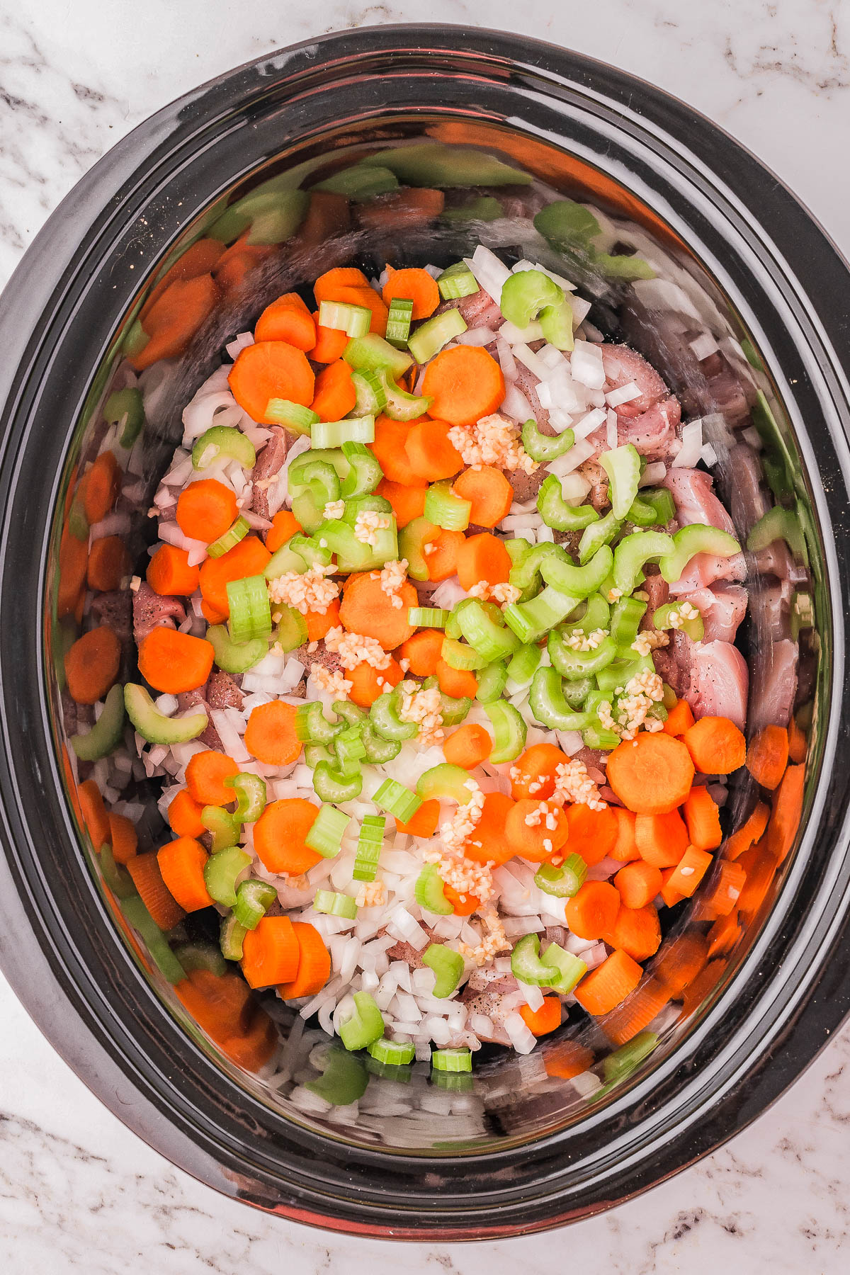 Top view of a slow cooker filled with chopped carrots, celery, onions, garlic, and pieces of meat on a marble countertop.