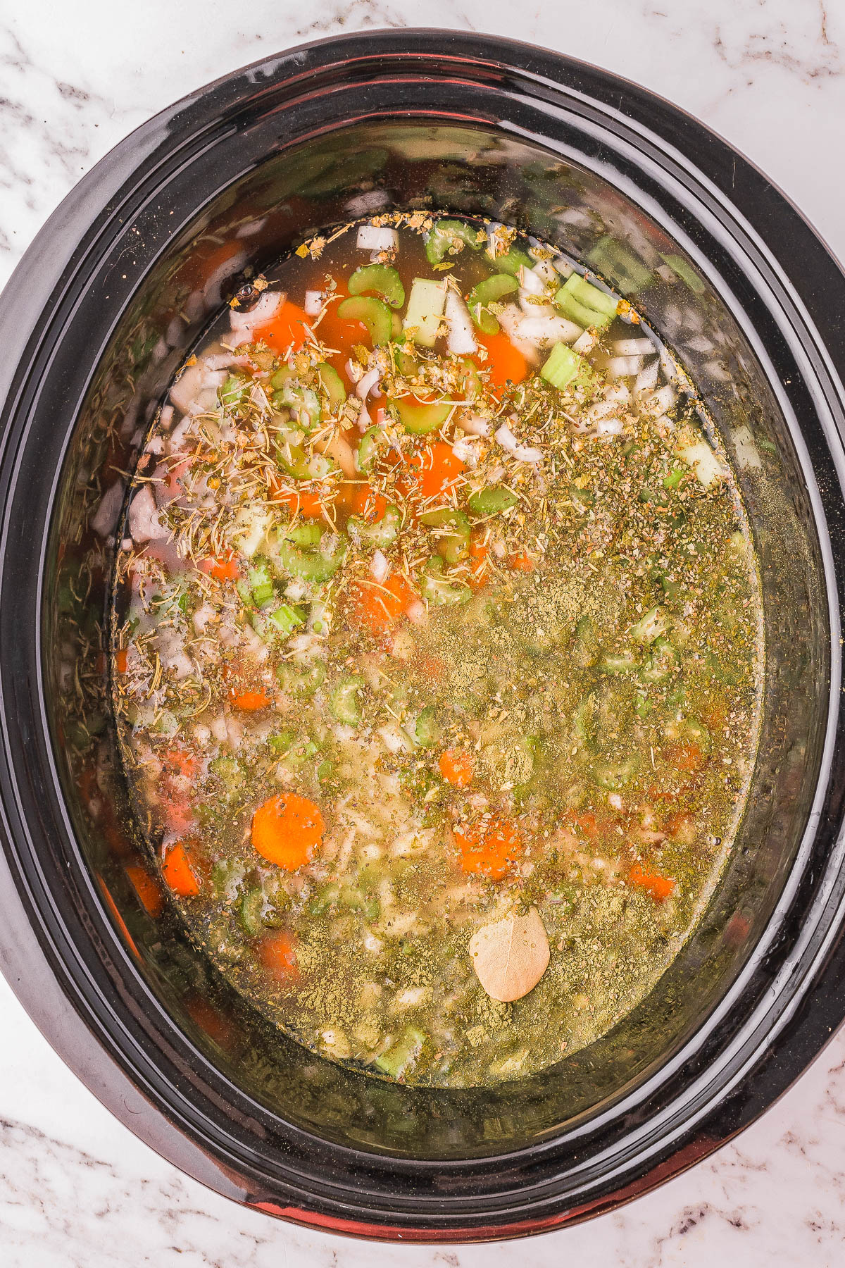 Slow cooker filled with chopped vegetables, herbs, and broth, preparing to be cooked.