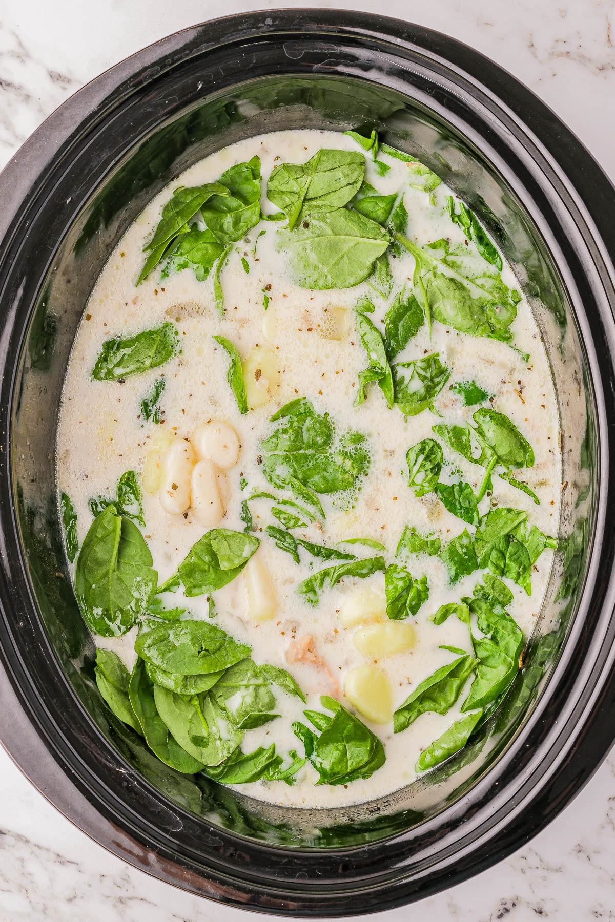 A slow cooker with creamy soup containing spinach, potatoes, and beans on a marble countertop.