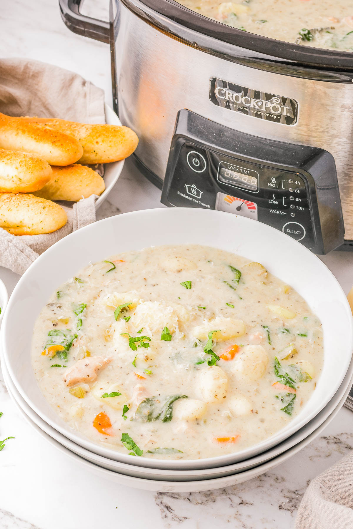 Bowl of creamy soup with vegetables and herbs in front of a Crock-Pot, next to a plate of breadsticks.