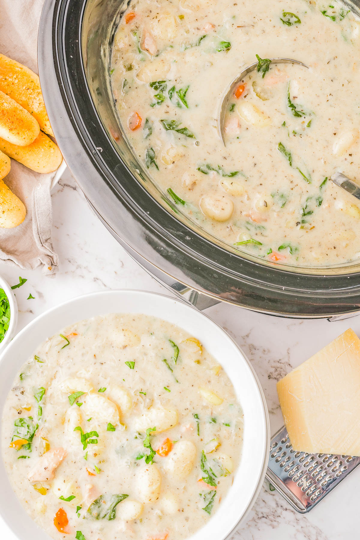 A bowl and slow cooker filled with creamy soup containing vegetables and gnocchi. Breadsticks and a block of cheese are on the side.