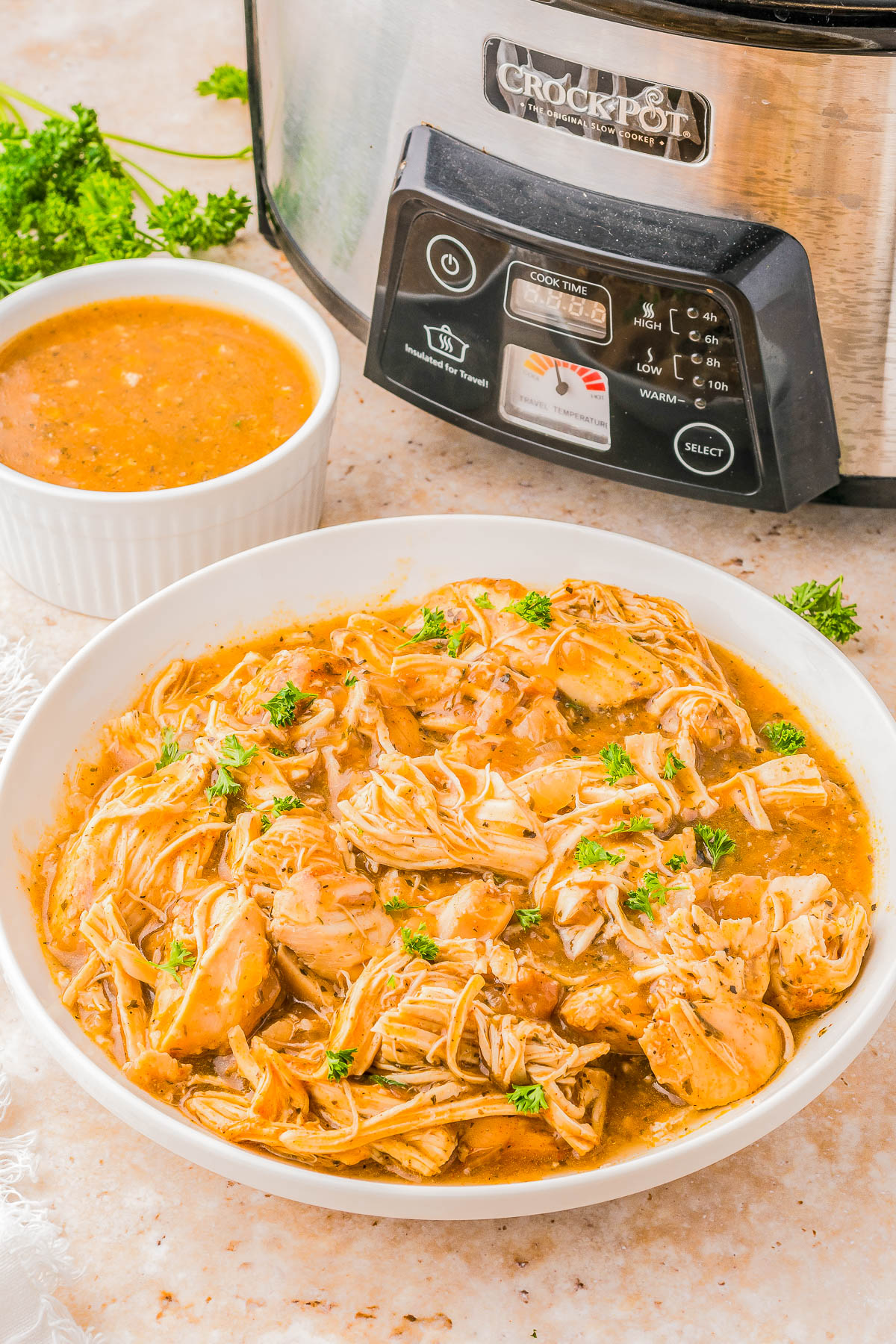 A plate of shredded chicken in sauce garnished with herbs, next to a bowl of sauce and a slow cooker in the background.