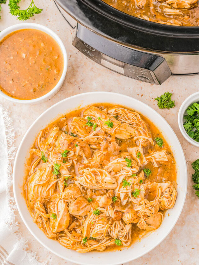 A bowl of shredded chicken in sauce, garnished with herbs, next to a slow cooker and a small bowl of extra sauce on a marble surface.