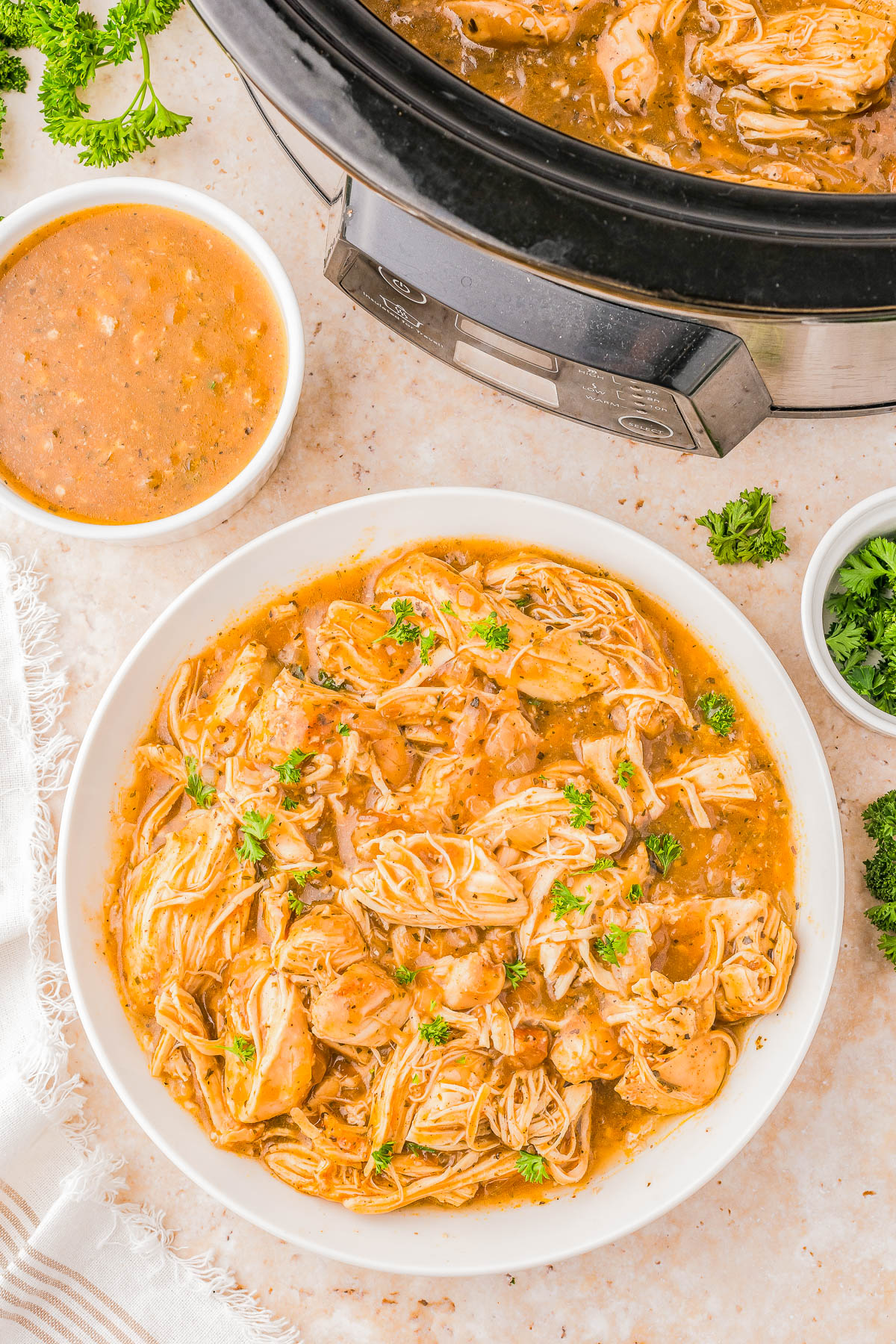 A bowl of shredded chicken in sauce, garnished with herbs, next to a slow cooker and a small bowl of extra sauce on a marble surface.