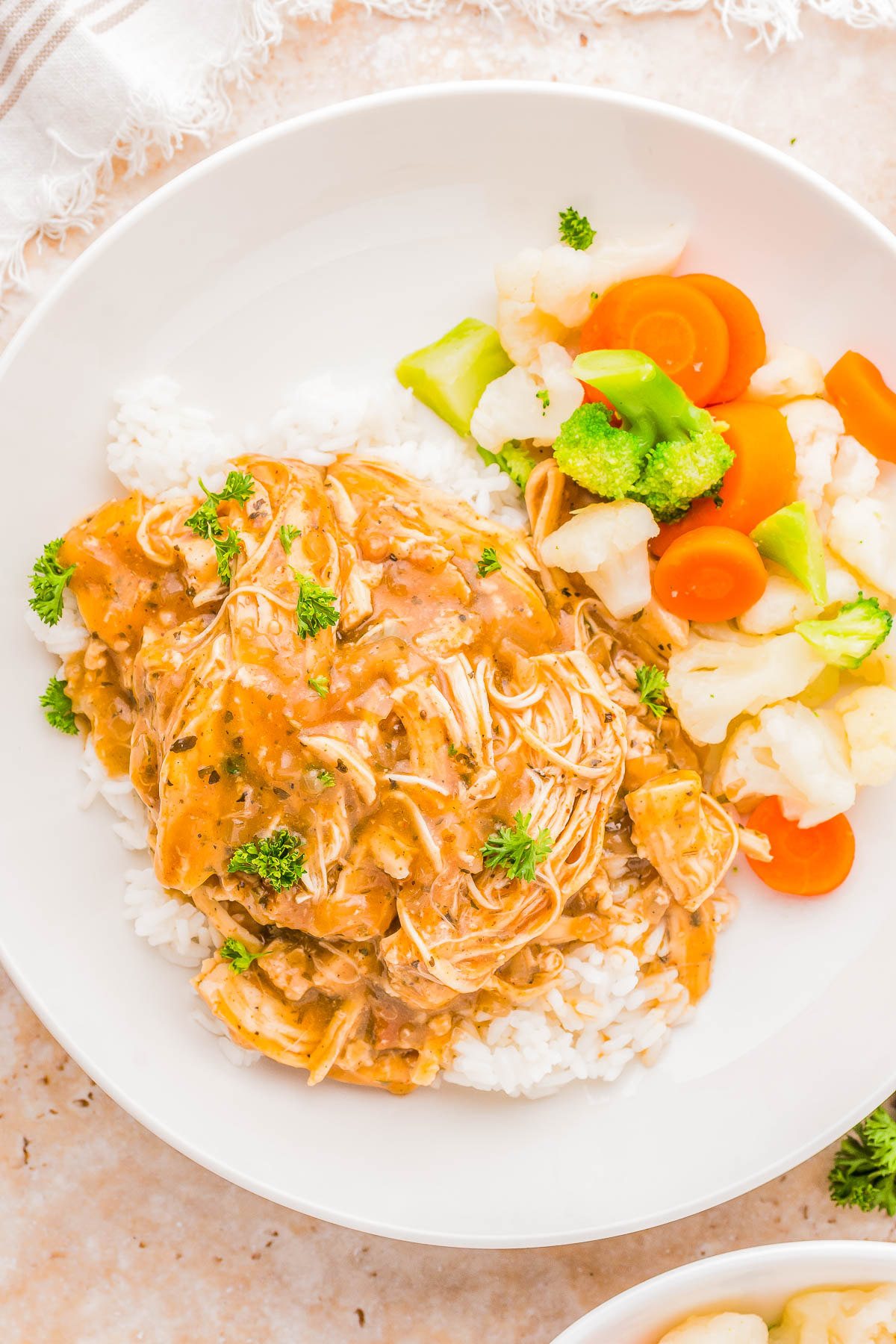 Plate of shredded chicken in gravy over rice, garnished with parsley, served with steamed broccoli, carrots, and cauliflower on the side.