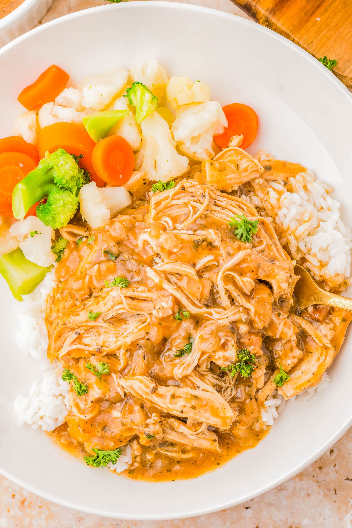 A plate of shredded chicken in gravy served over rice with a side of steamed vegetables, including broccoli, carrots, and cauliflower.