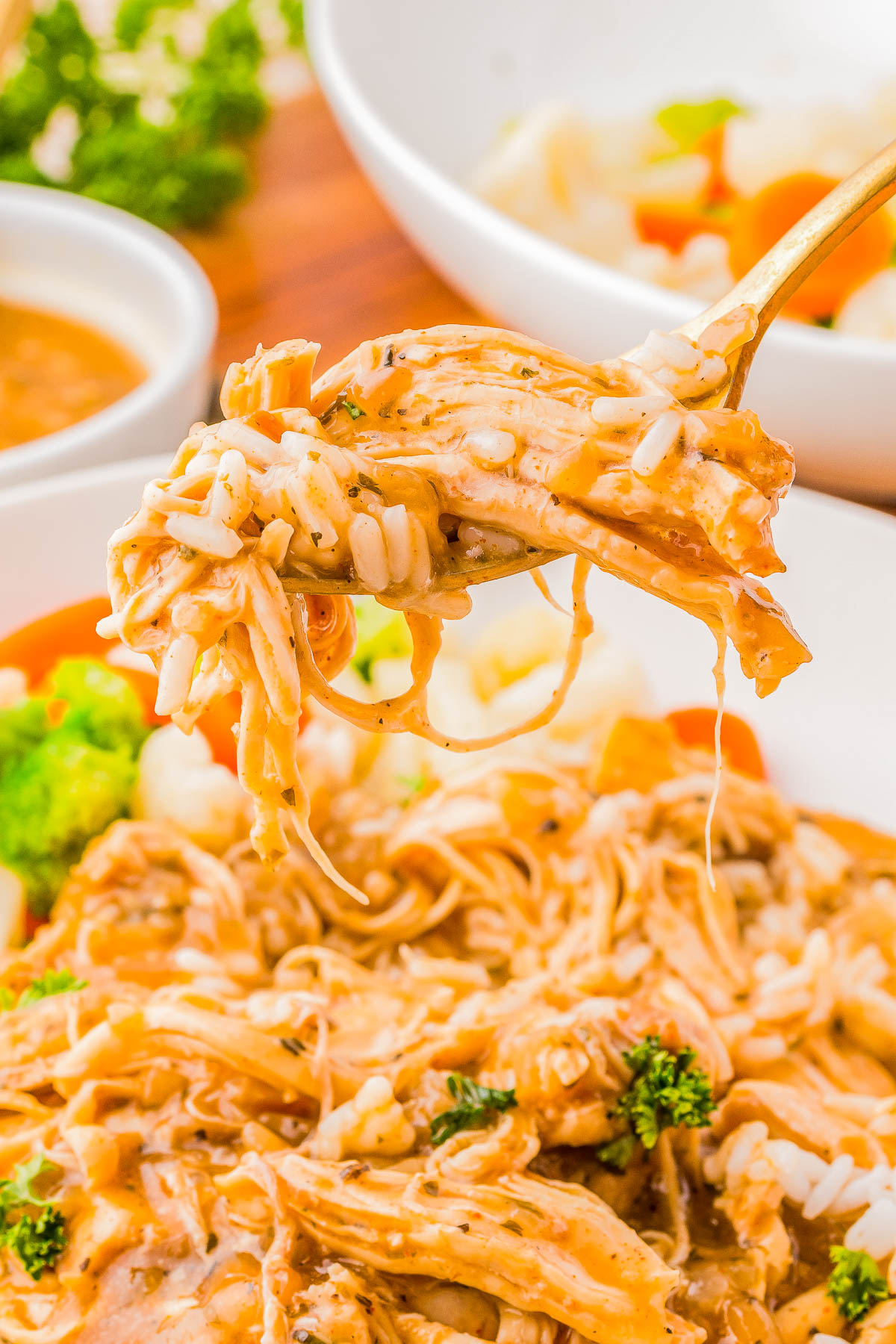 Close-up of a fork holding shredded chicken with rice, carrots, and broccoli in a creamy sauce, over a plate of the same dish. Bowls of sauce and vegetables are visible in the background.