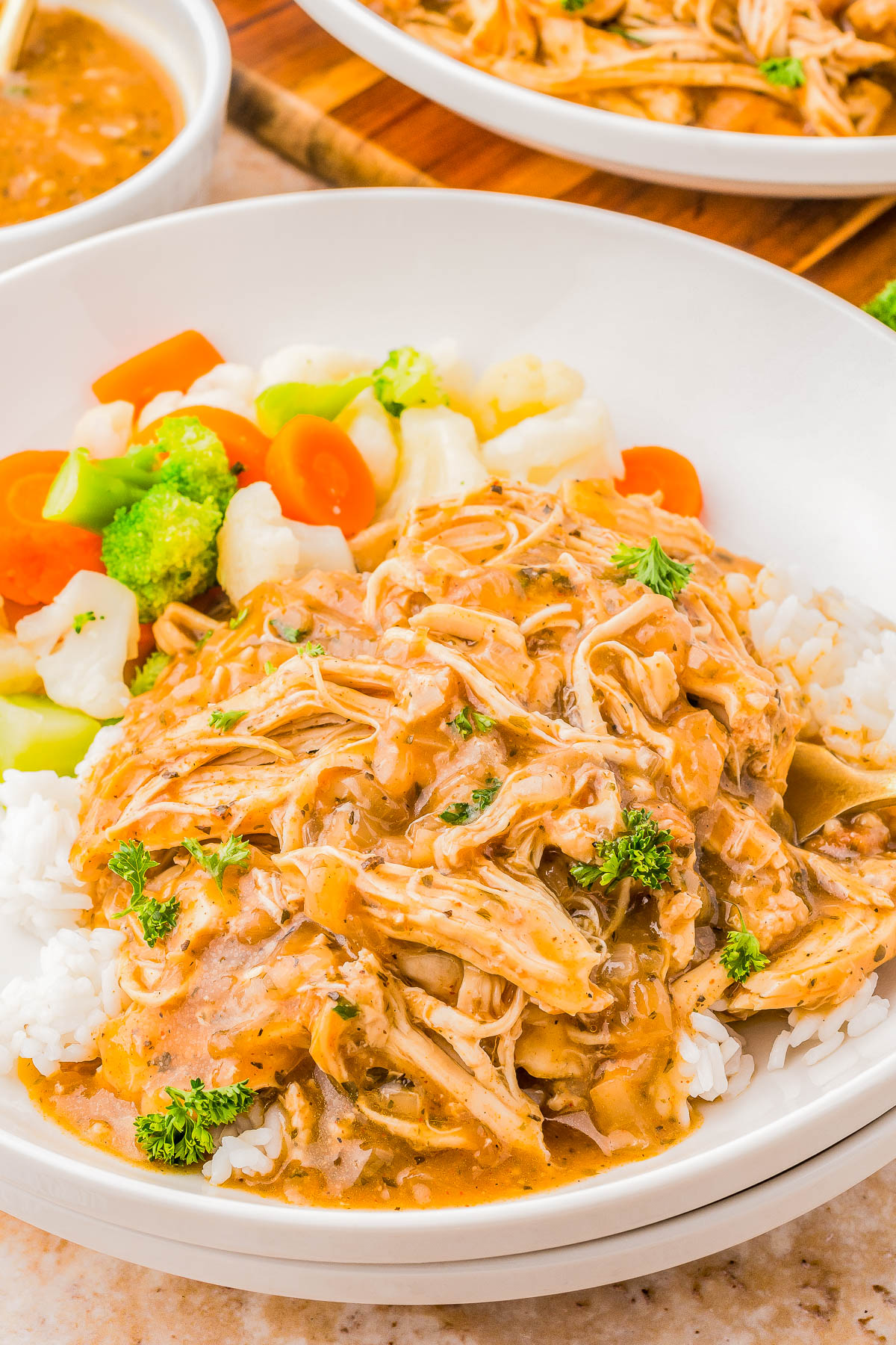 A bowl of shredded chicken in brown sauce over rice, garnished with parsley. Served with mixed steamed vegetables including broccoli, carrots, and cauliflower.