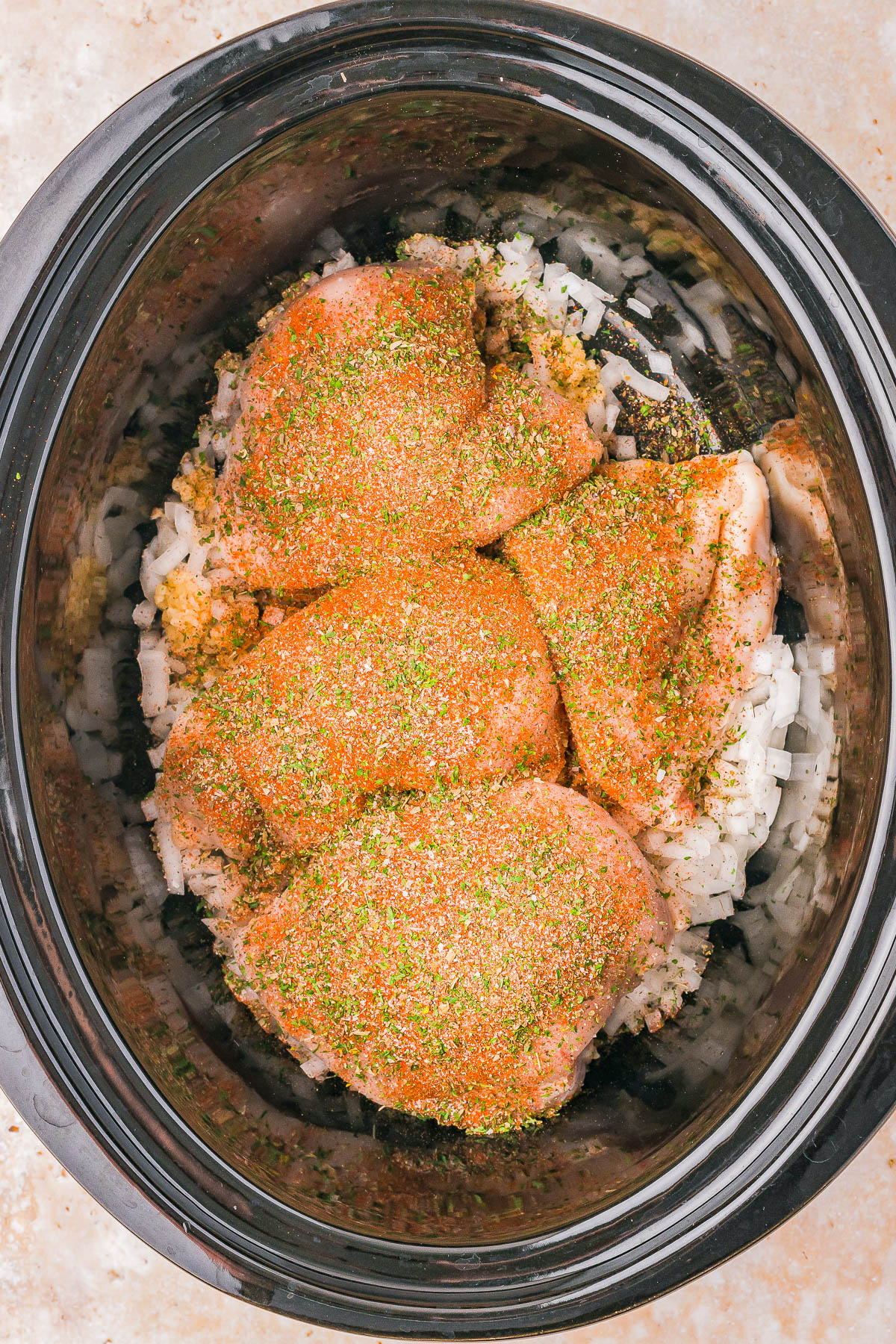 Chicken pieces seasoned with spices sit on top of chopped onions in a slow cooker.