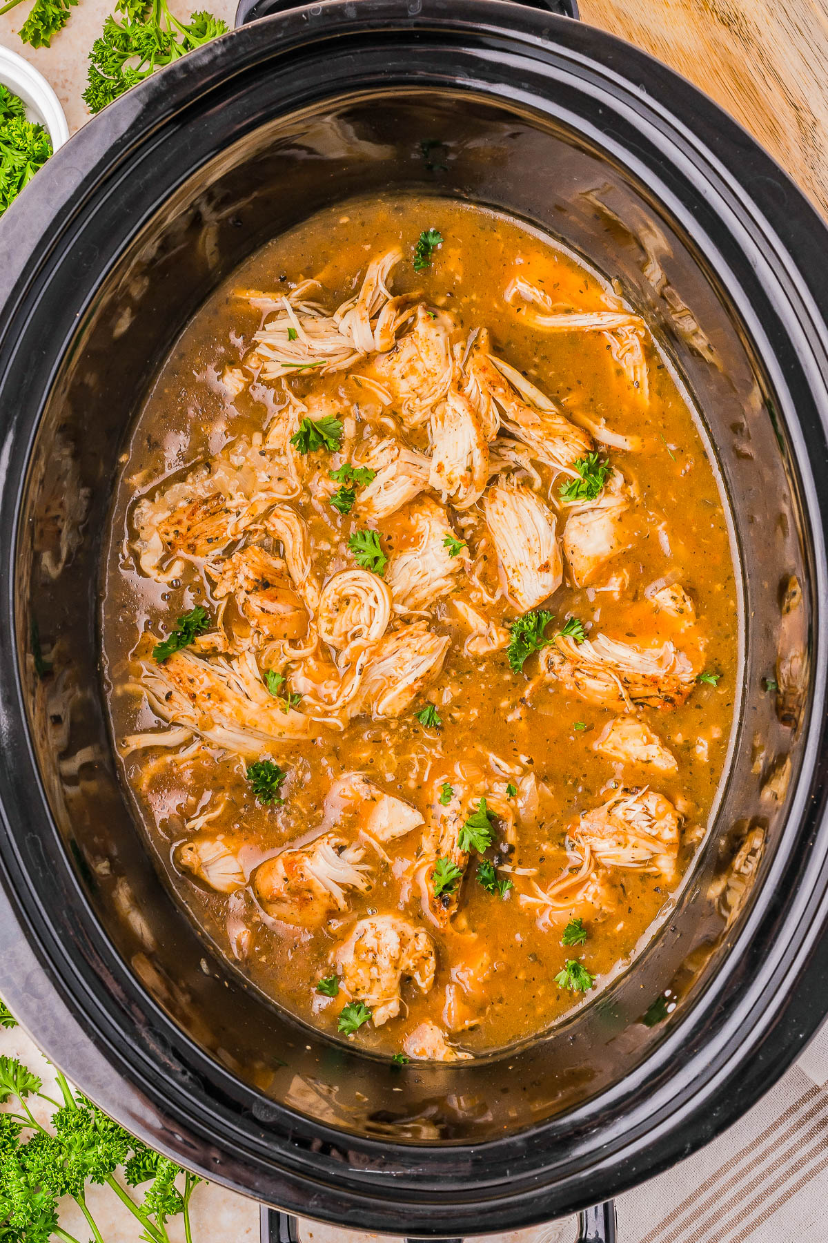 Shredded chicken in a slow cooker with a savory sauce, garnished with chopped parsley.