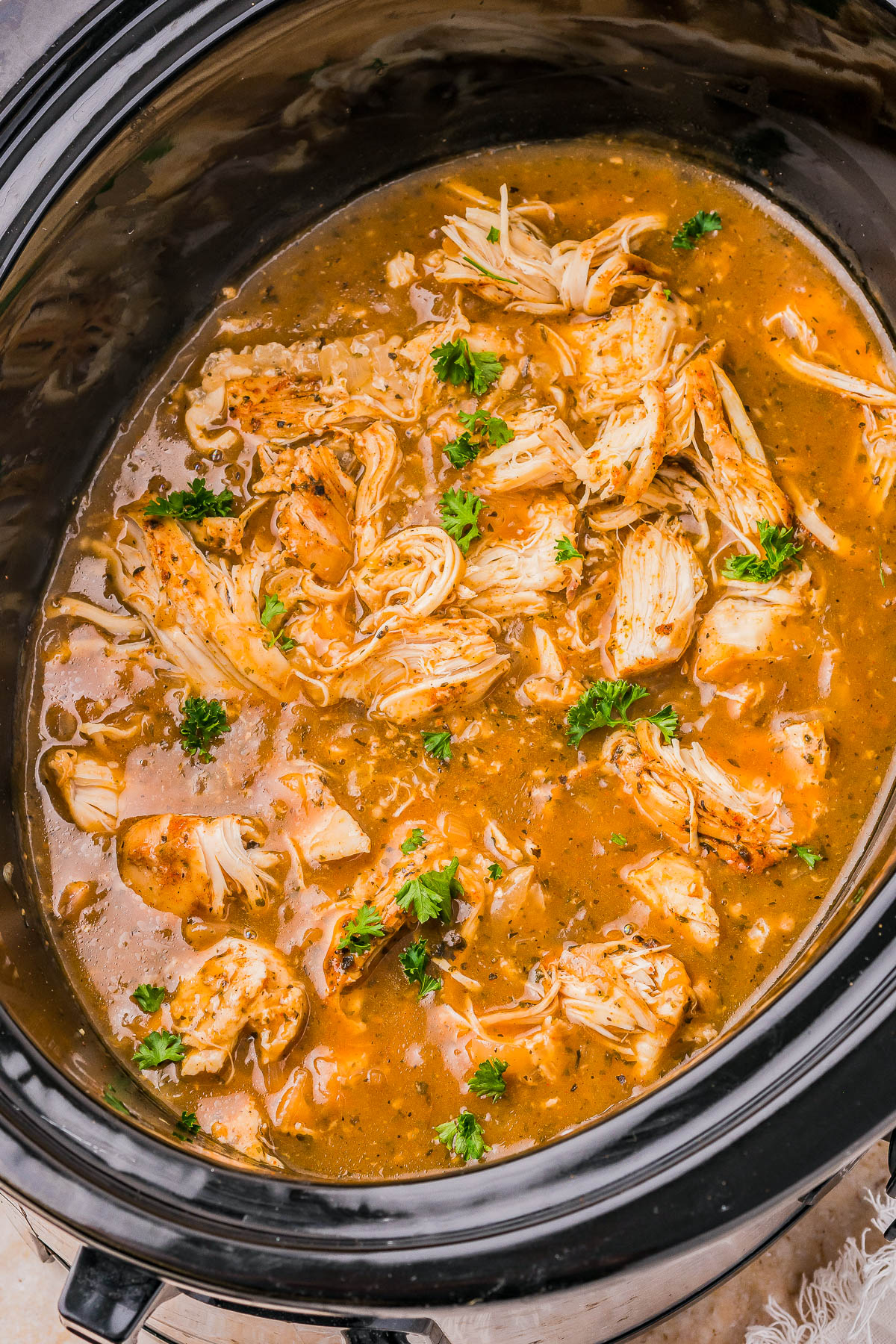 Shredded chicken in a slow cooker with a brown gravy, garnished with parsley.