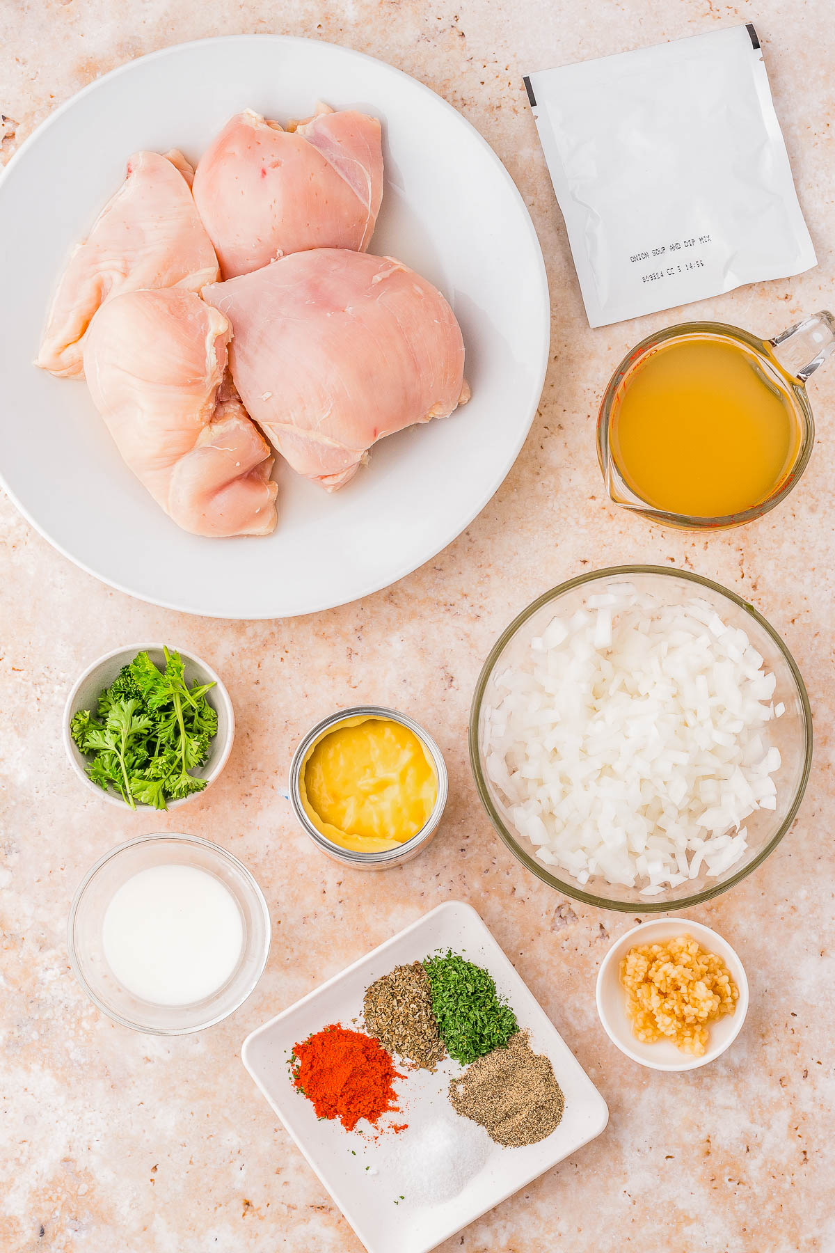 Raw chicken breasts on a plate surrounded by bowls of chopped onions, garlic, herbs, spices, cream, pineapple, broth, and a seasoning packet on a marble countertop.