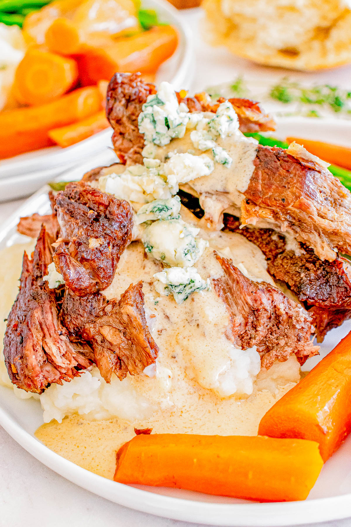 Plate of braised beef on mashed potatoes, topped with blue cheese sauce, surrounded by cooked carrots and asparagus.