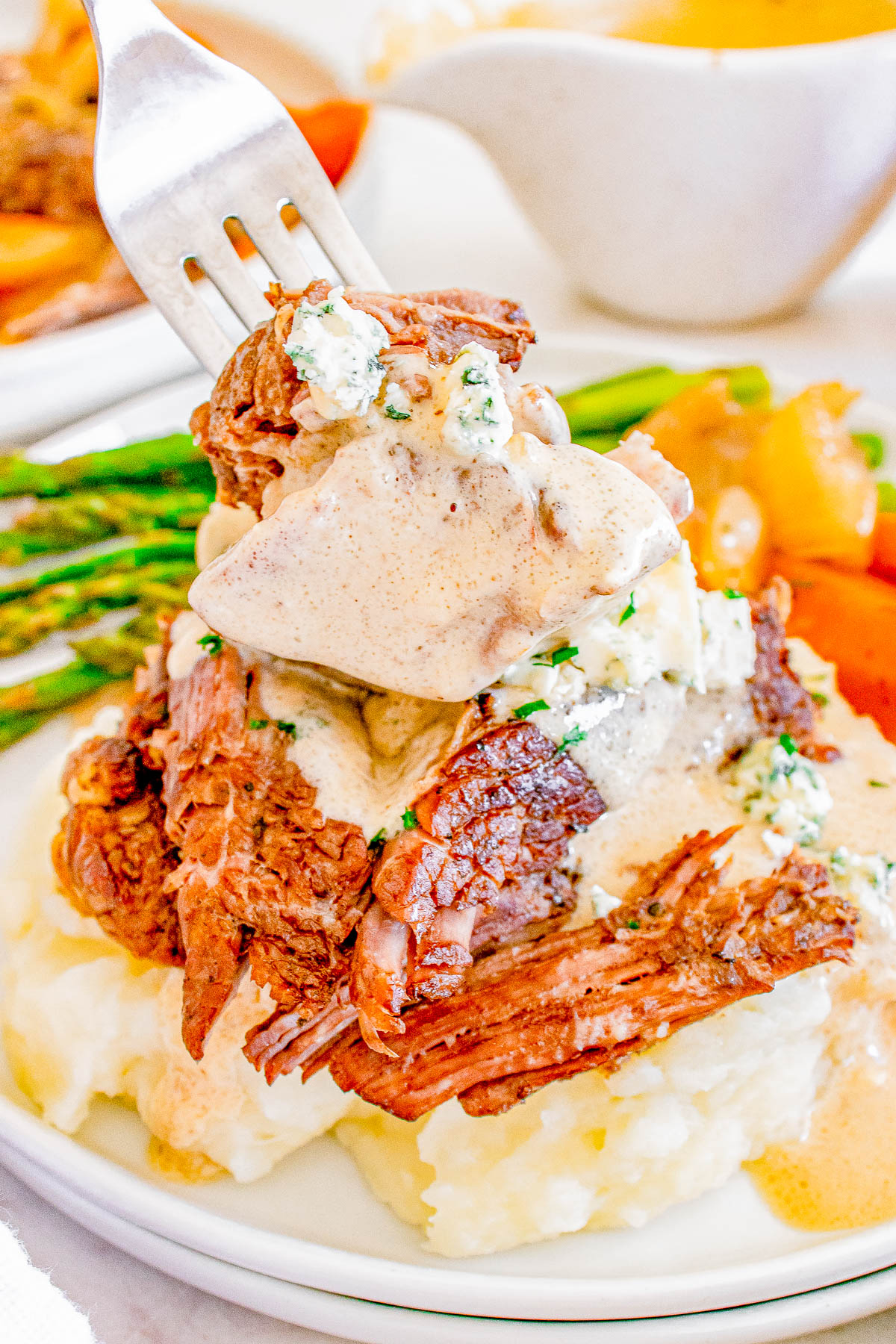 A fork holds a piece of beef with creamy sauce, placed on mashed potatoes. Asparagus and carrots are visible in the background.