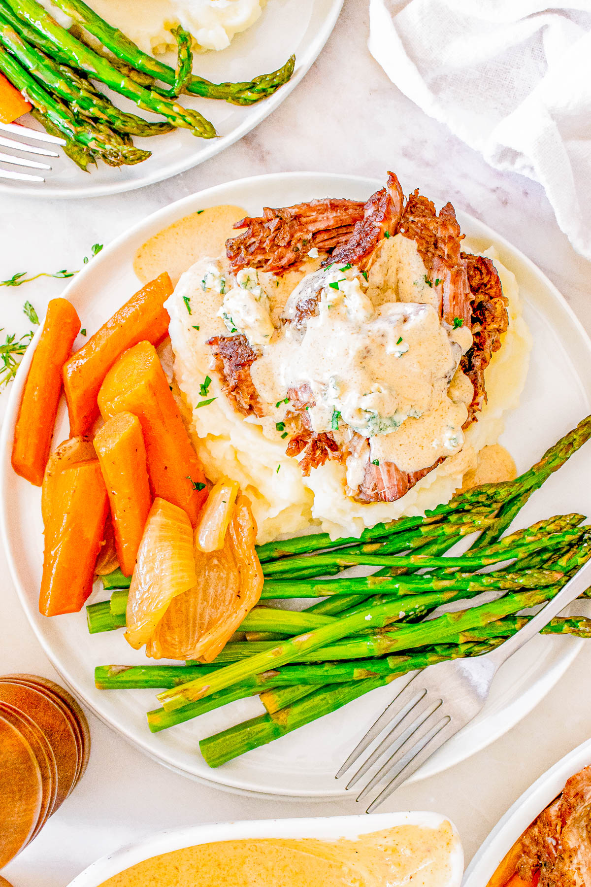 A plate of mashed potatoes topped with shredded meat and sauce, served with roasted carrots, onions, and asparagus.