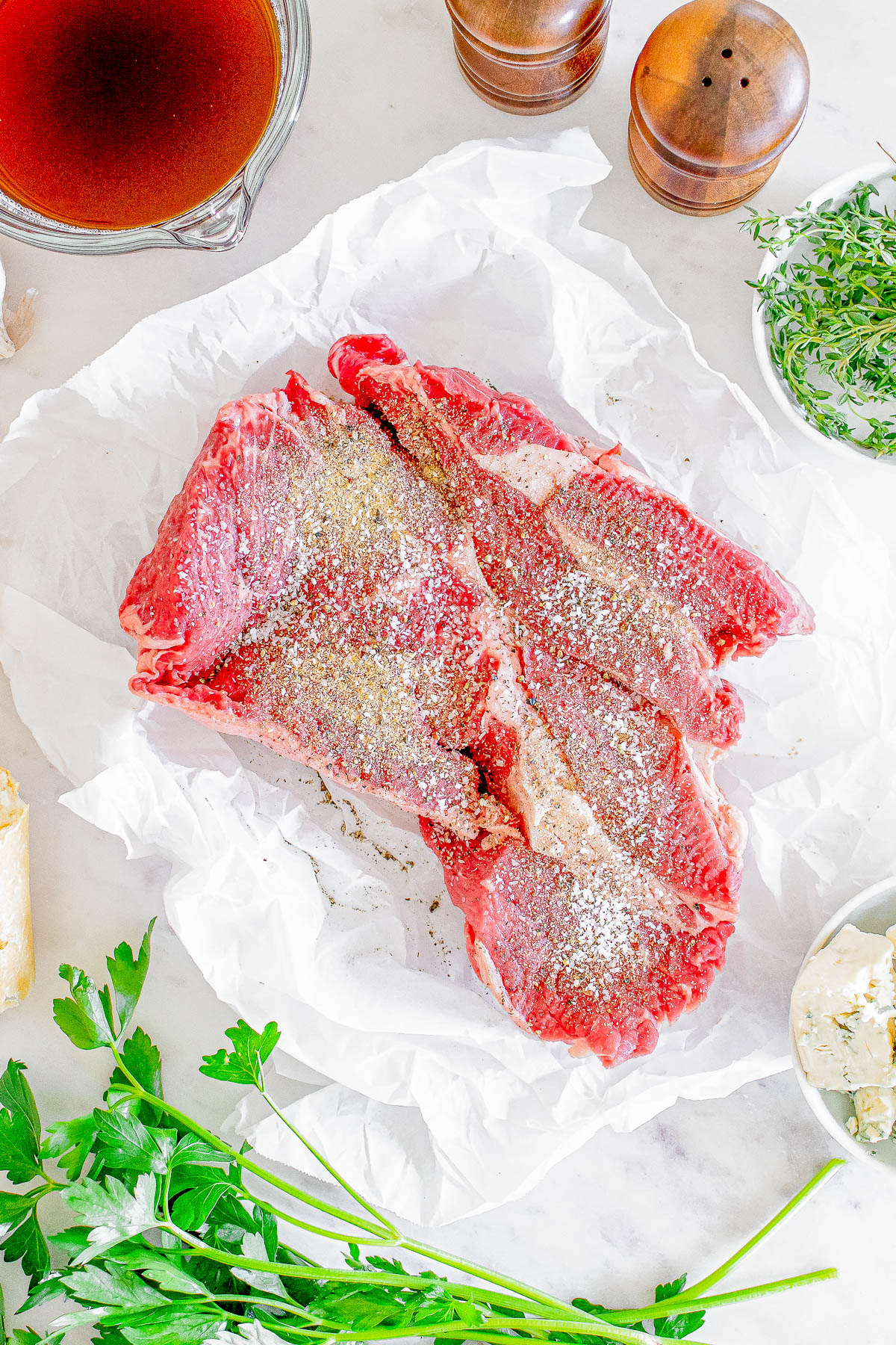 Raw beef steak on parchment paper, sprinkled with salt and pepper, surrounded by herbs, a gravy boat, and pepper mills on a white surface.