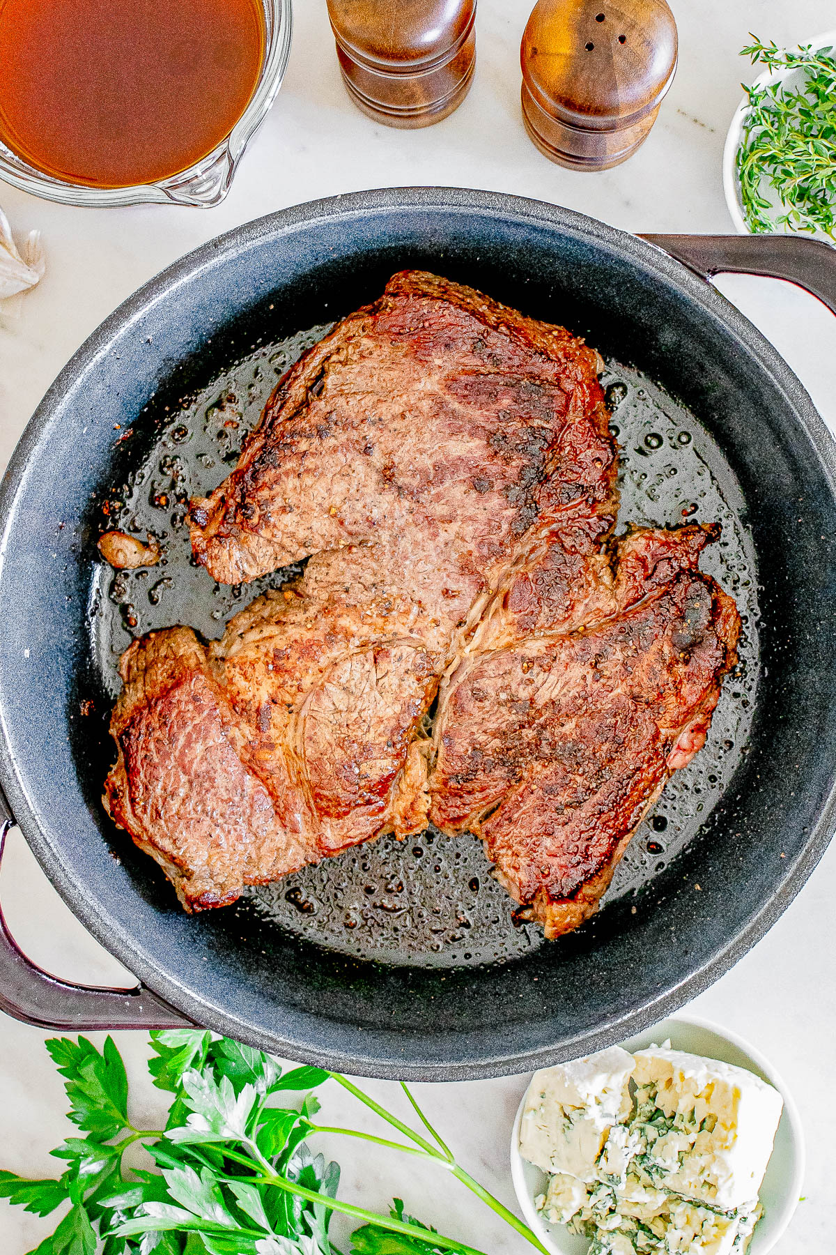 Two cooked steaks in a black pan with fresh herbs, blue cheese, and a bowl of sauce nearby on a white surface.